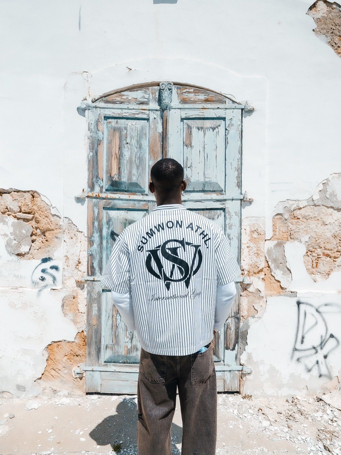 Man wearing Sumwon Athl striped long sleeve shirt, standing before a weathered blue door on a rustic wall.