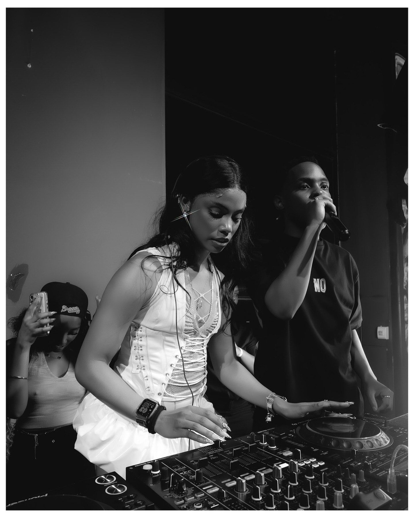 Black and white photo of a female DJ mixing at turntables while a male rapper performs with a microphone at a music event.