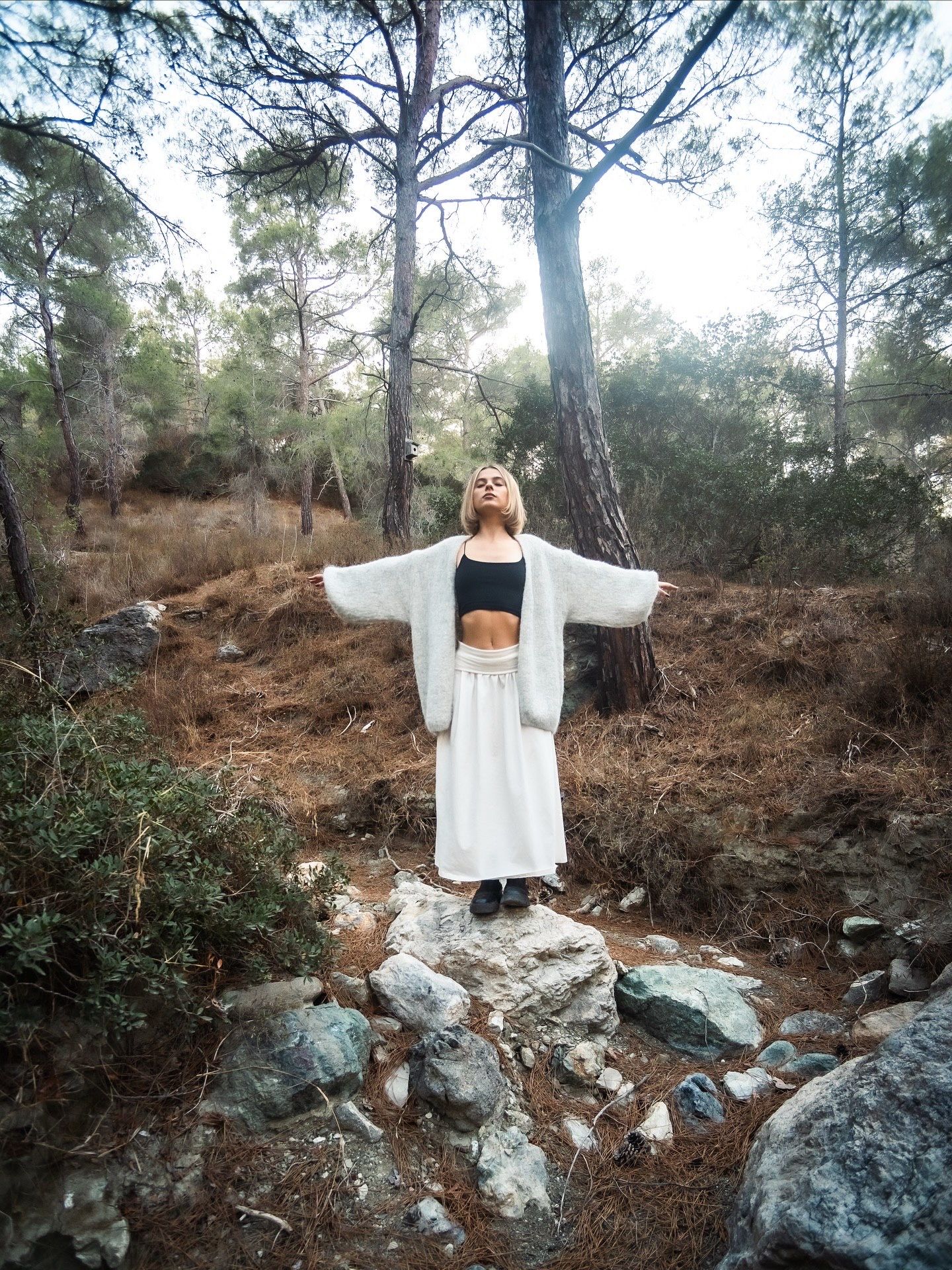 Woman in white kimono and skirt stands arms outstretched in a rocky forest, looking up toward the sky.
