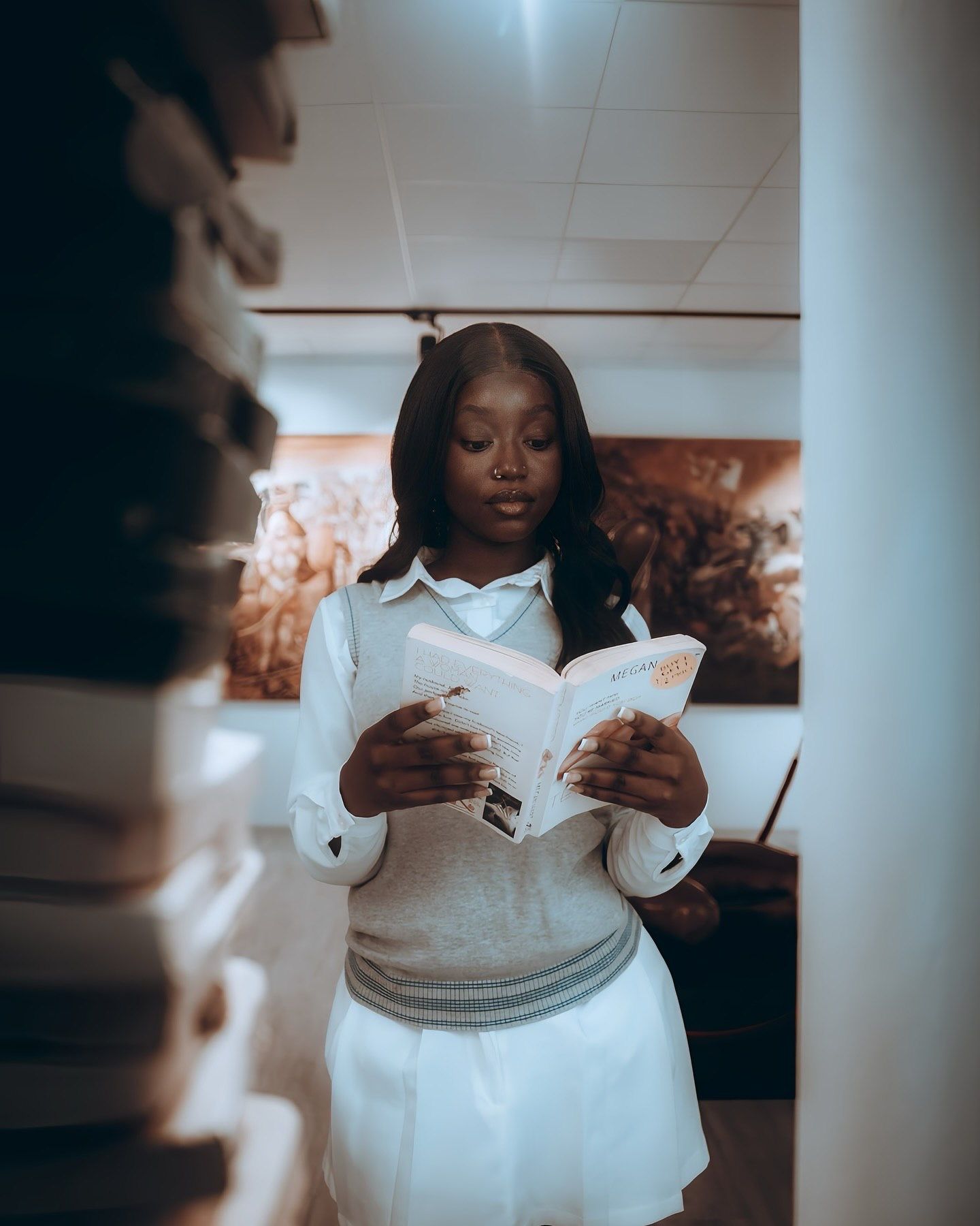 Young woman in white sweater and skirt reads a book surrounded by stacked books in a library setting.