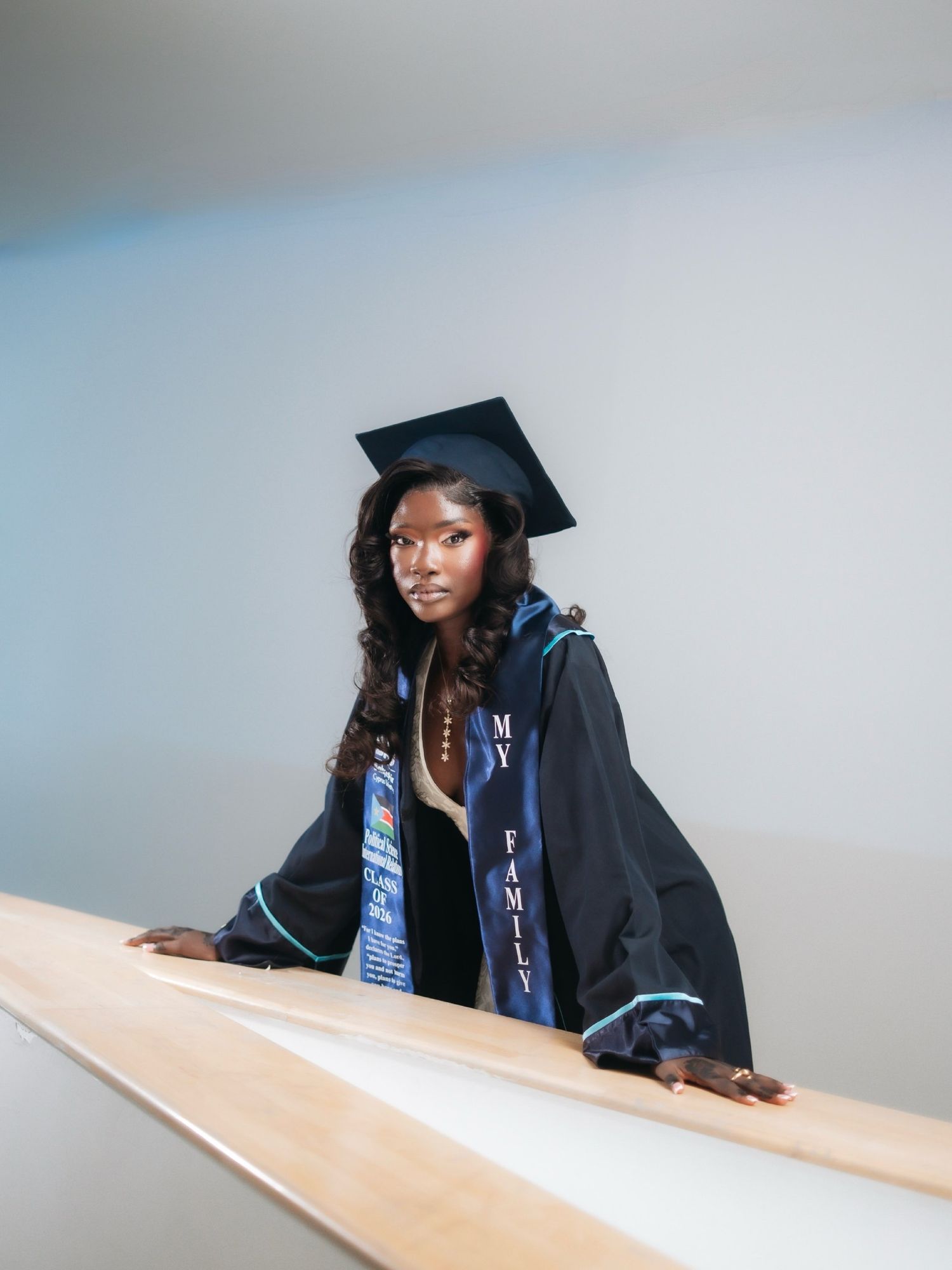 Graduate in cap and gown with 'My Family' stole poses on a staircase railing indoors.