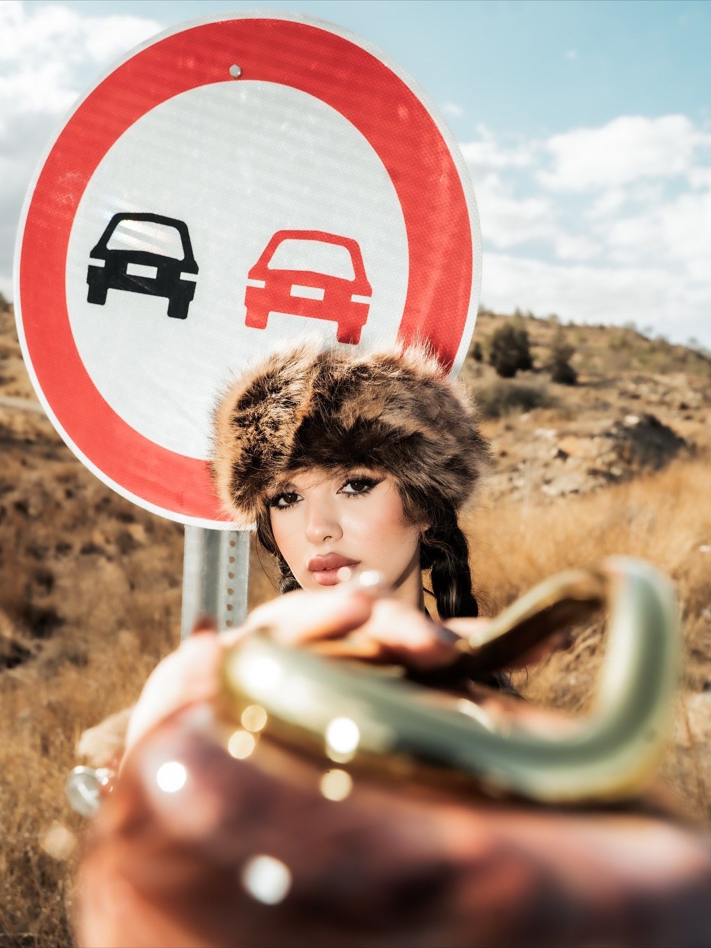 Woman in fur hat poses near a no-overtaking road sign in a rural landscape, holding a decorative object.