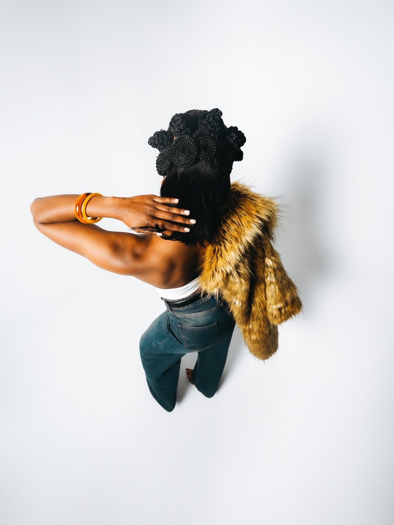 Overhead view of a woman in fur jacket and jeans with a black crochet headpiece against white wall.