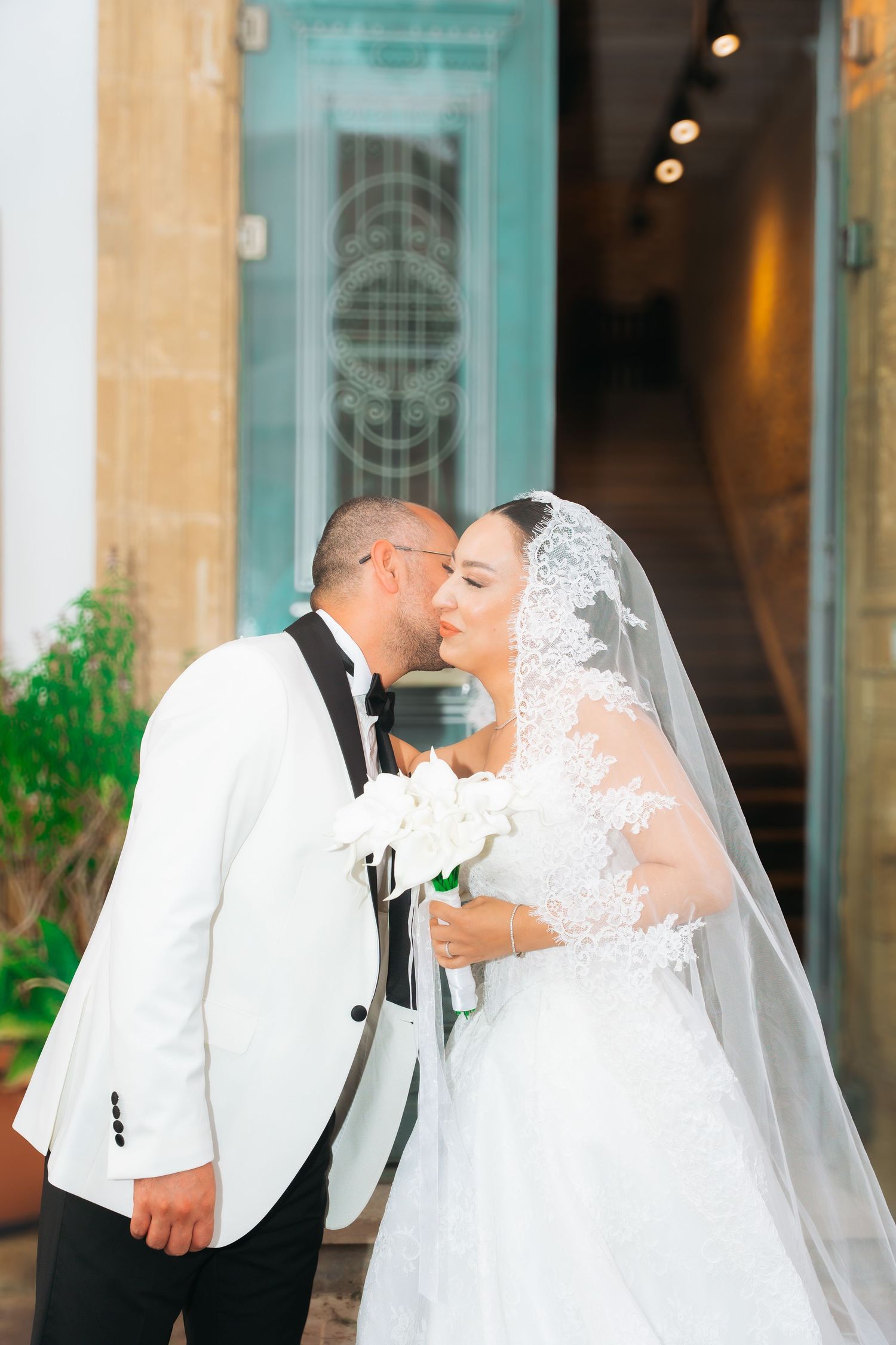 Bride and groom sharing a kiss outside a church, bride in lace veil holding white bouquet, groom in white tuxedo.