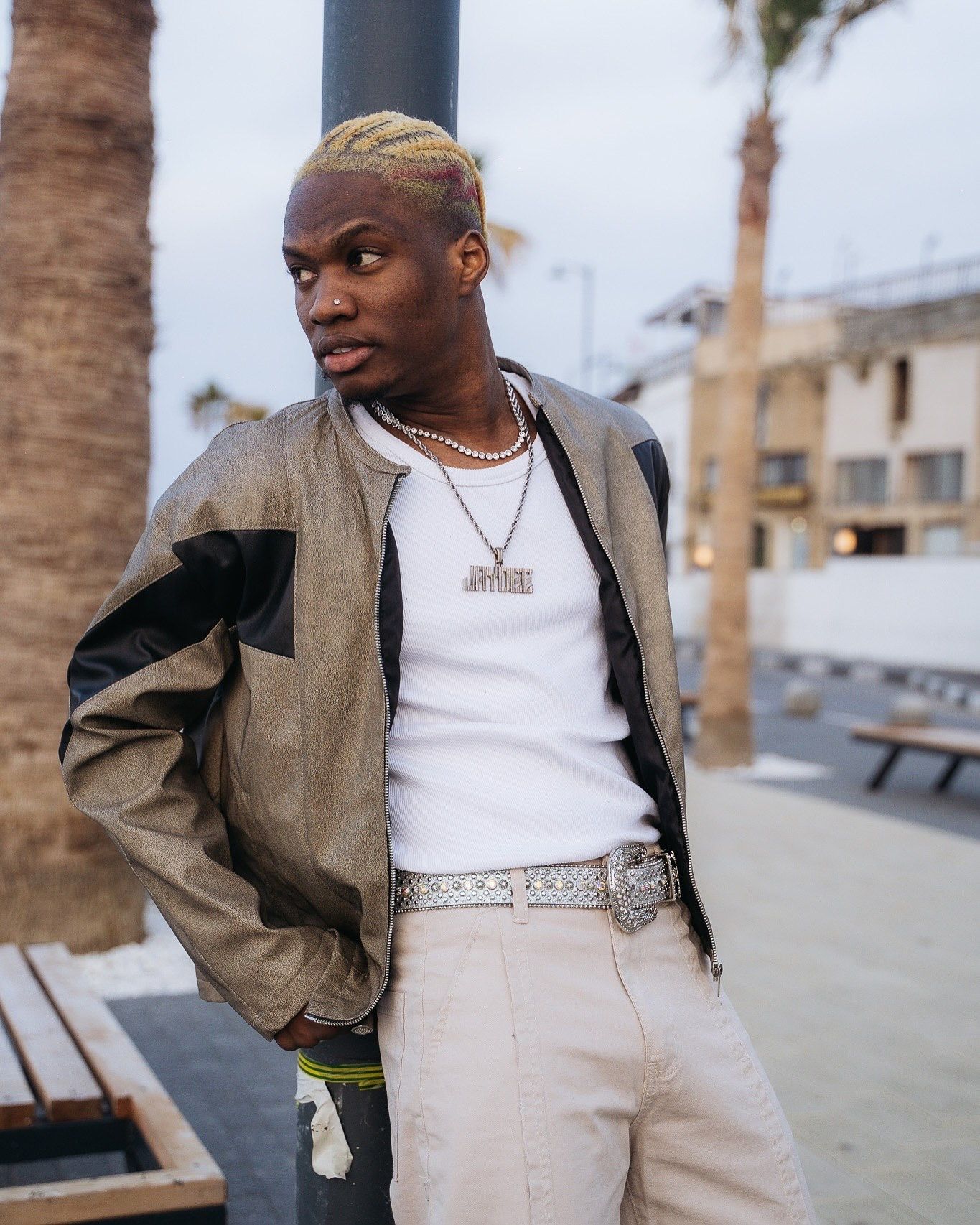 Young man with blonde-tipped hair wearing a leather jacket, white tee, chain necklace, and cream pants outdoors.