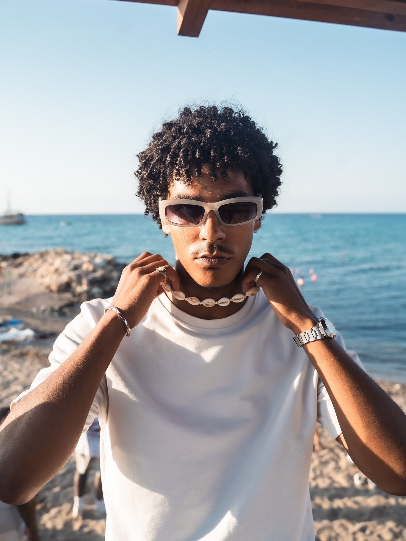 Young man with curly hair wearing white sunglasses and shell necklace at a sunny beach.