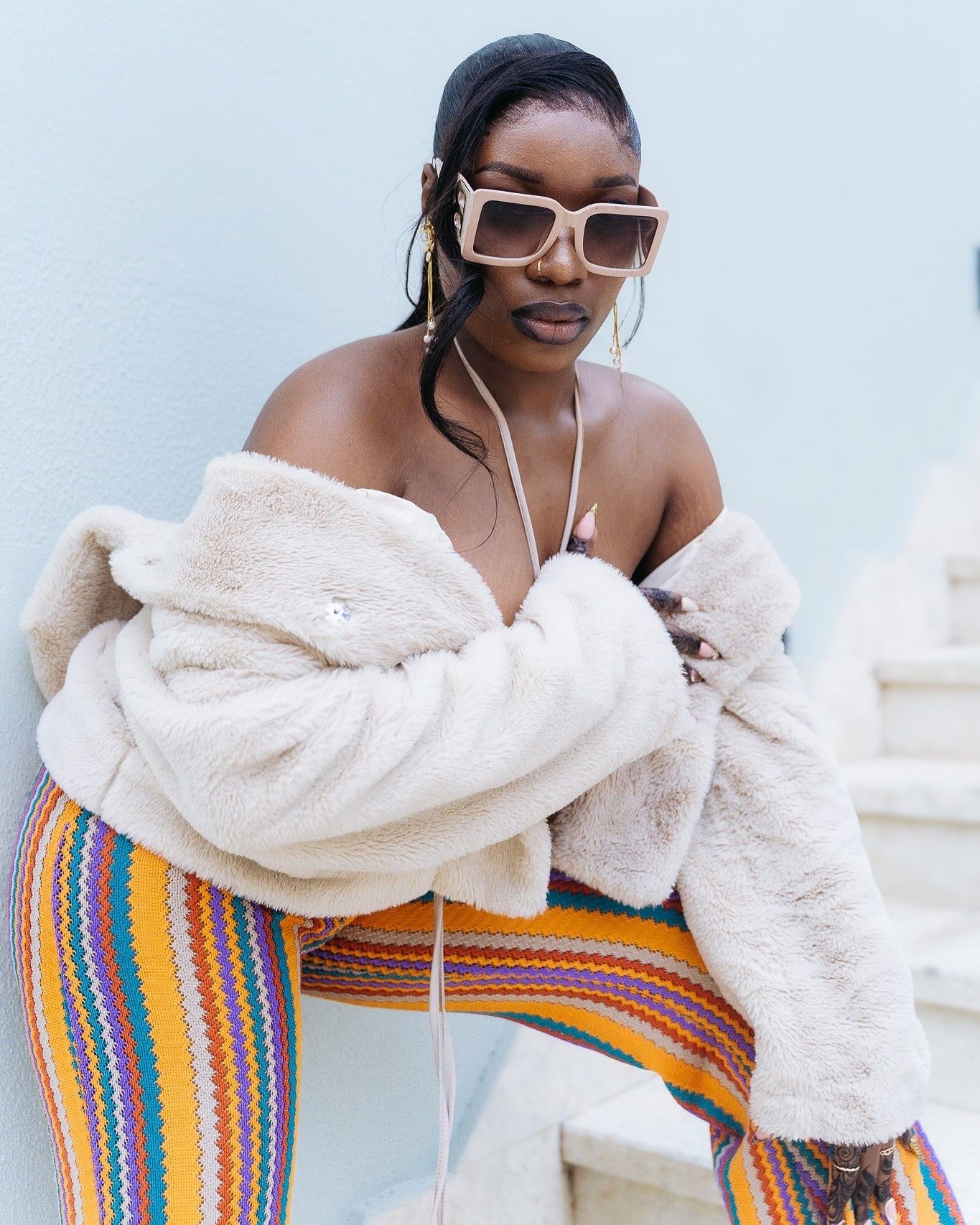 Woman in colorful striped pants, fluffy cream jacket, and oversized square sunglasses posing outdoors.