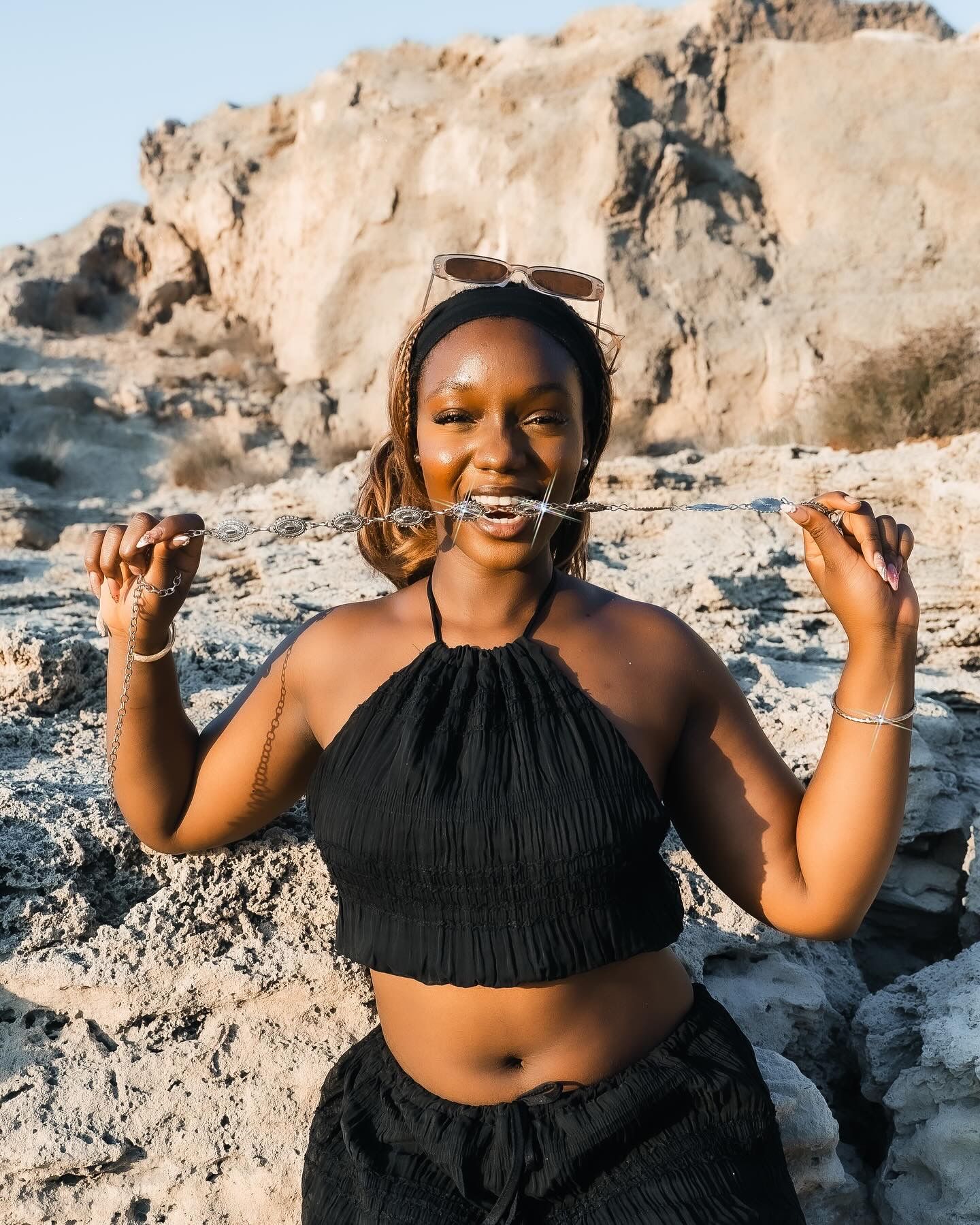 A smiling woman in a black crop top holds sunglasses in her mouth while posing among rocky desert terrain.