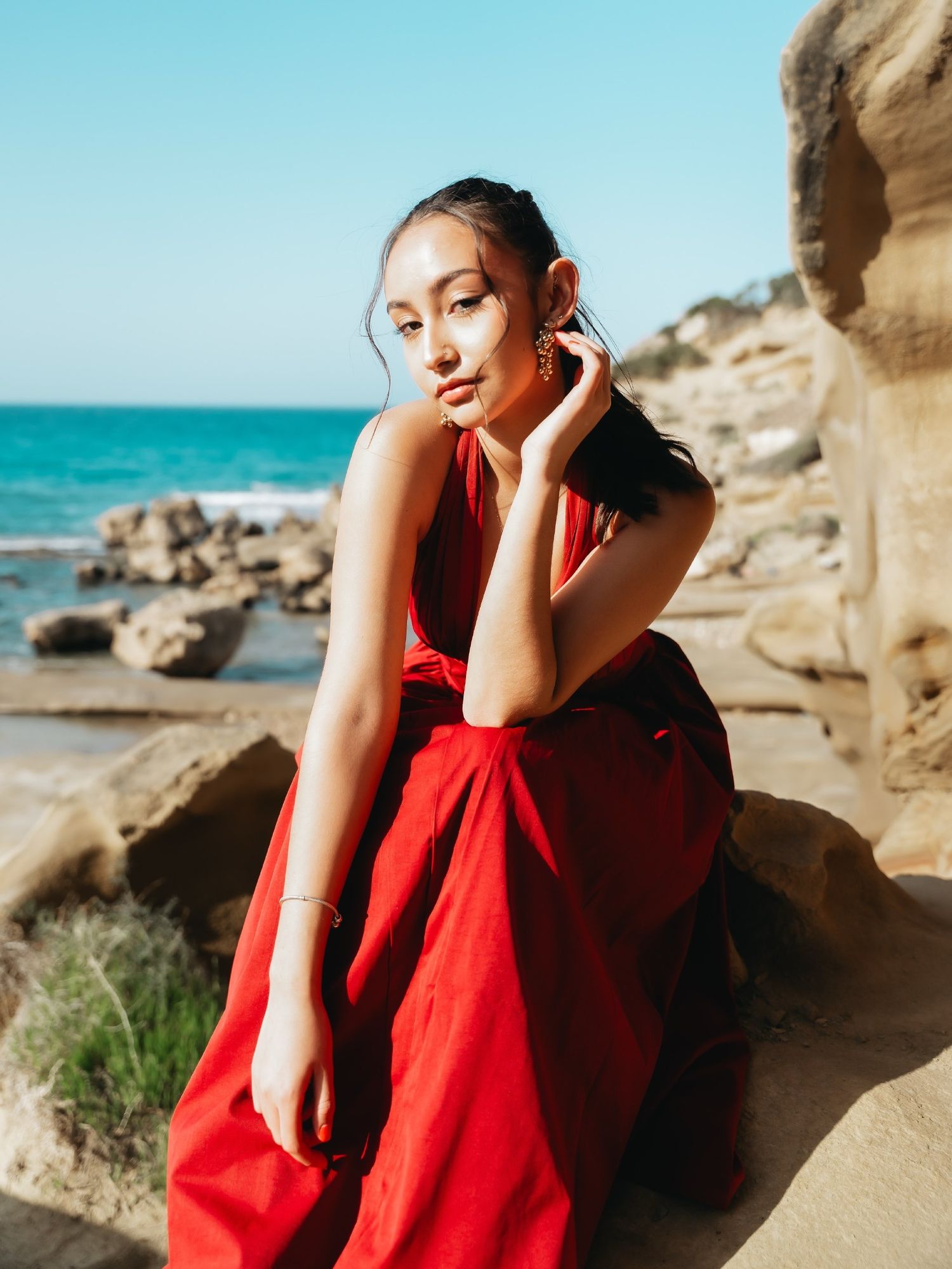 Woman in red dress sitting on coastal rocks with ocean view behind her.