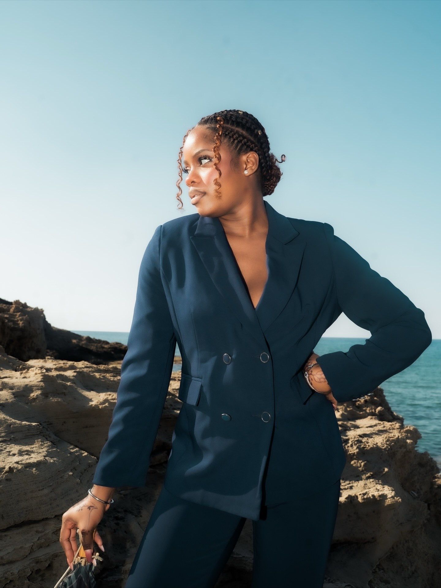 Woman in navy blue double-breasted blazer suit poses on rocky coastal cliffs with ocean backdrop.