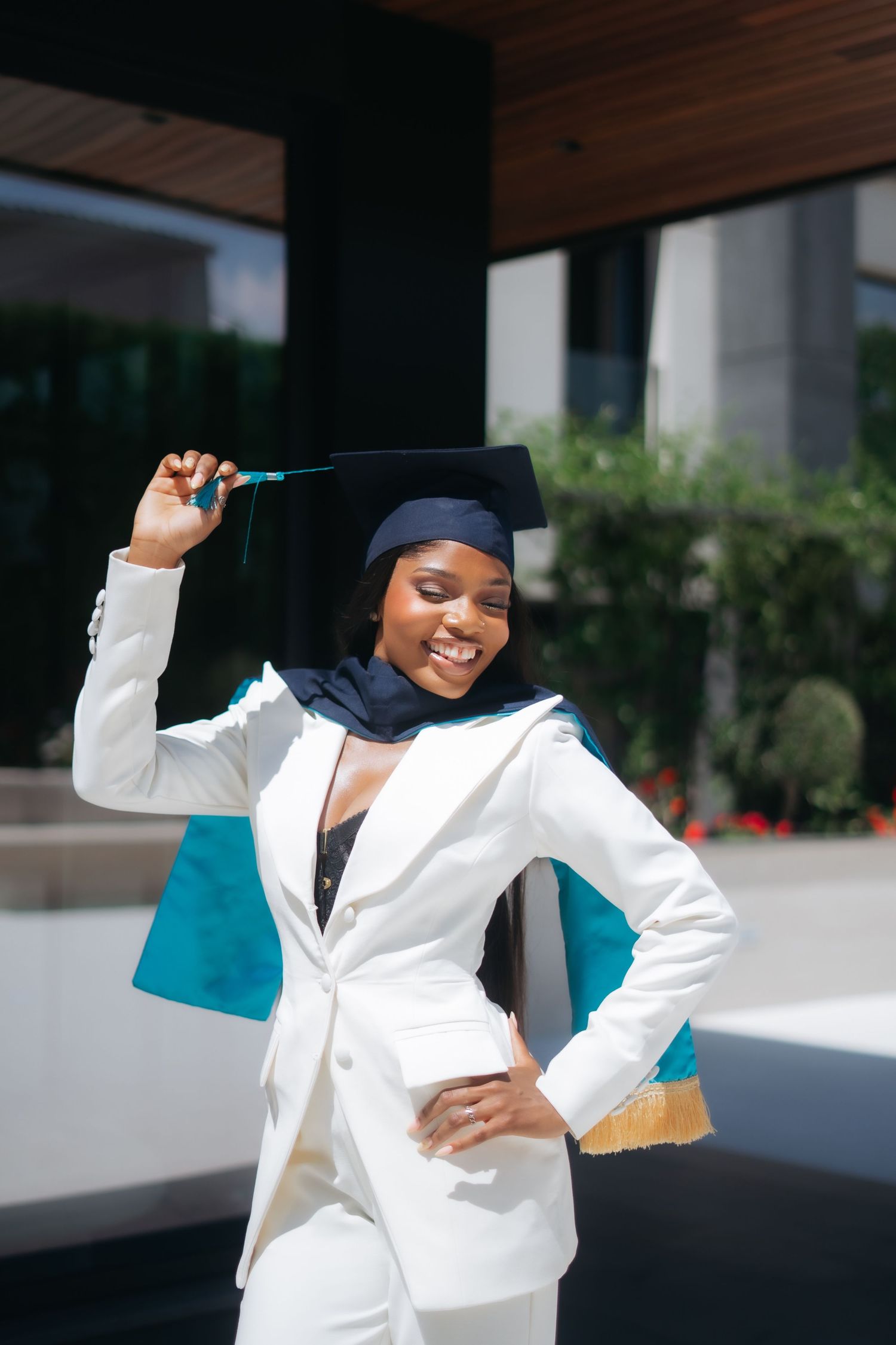 Smiling Black Muslim woman in hijab and white suit adjusts navy graduation cap outdoors.