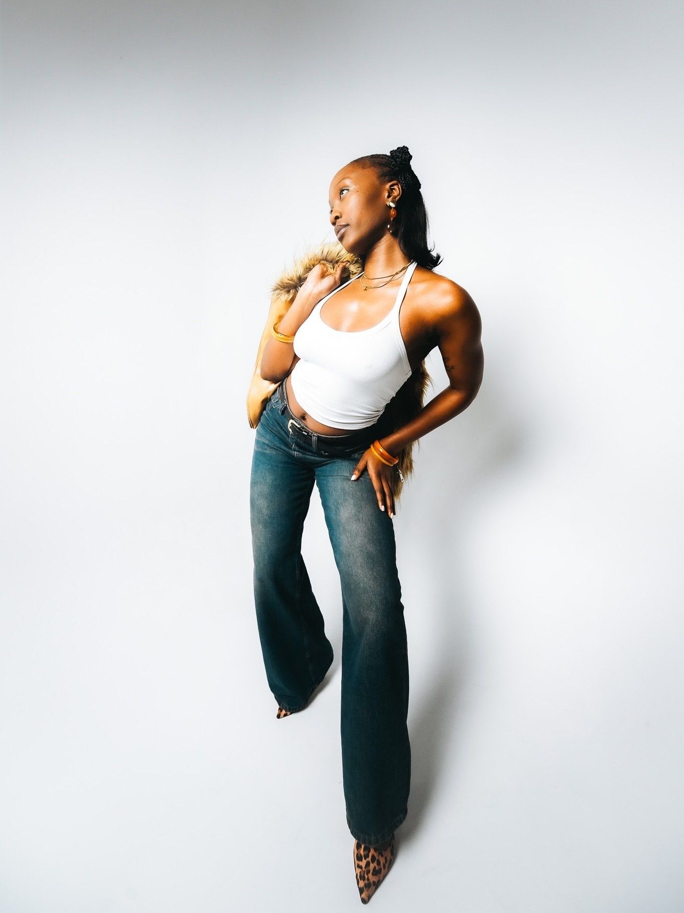 Tall Black woman in white halter top, flared jeans, and leopard heels posing against white background.