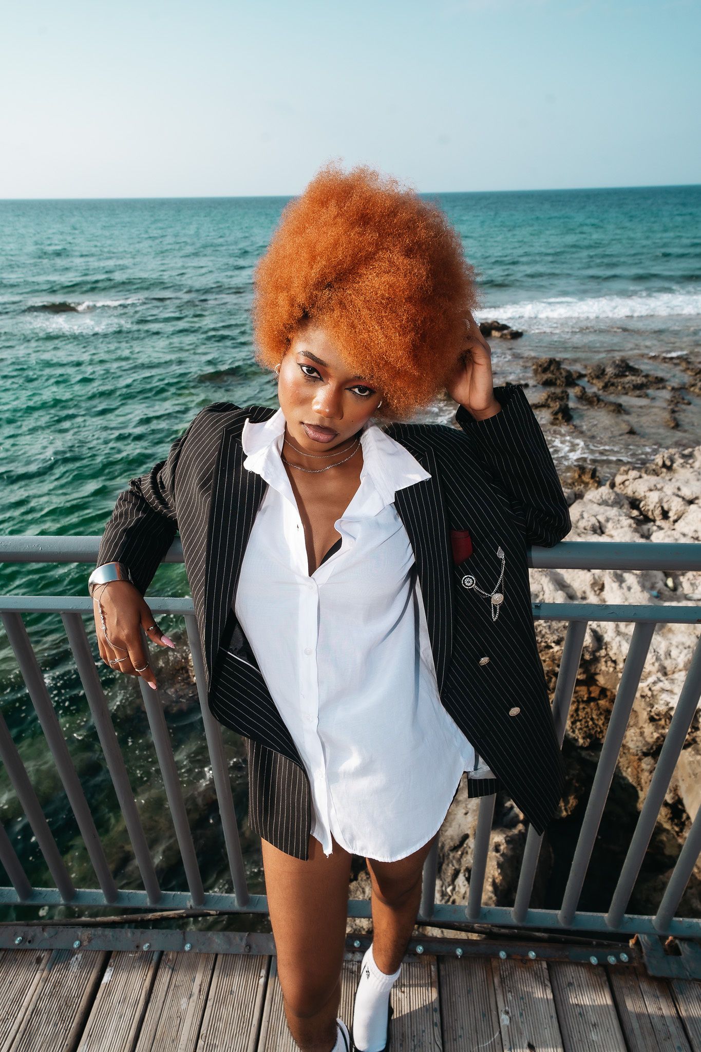Woman with voluminous red afro in oversized pinstripe blazer poses on rocky seaside railing.
