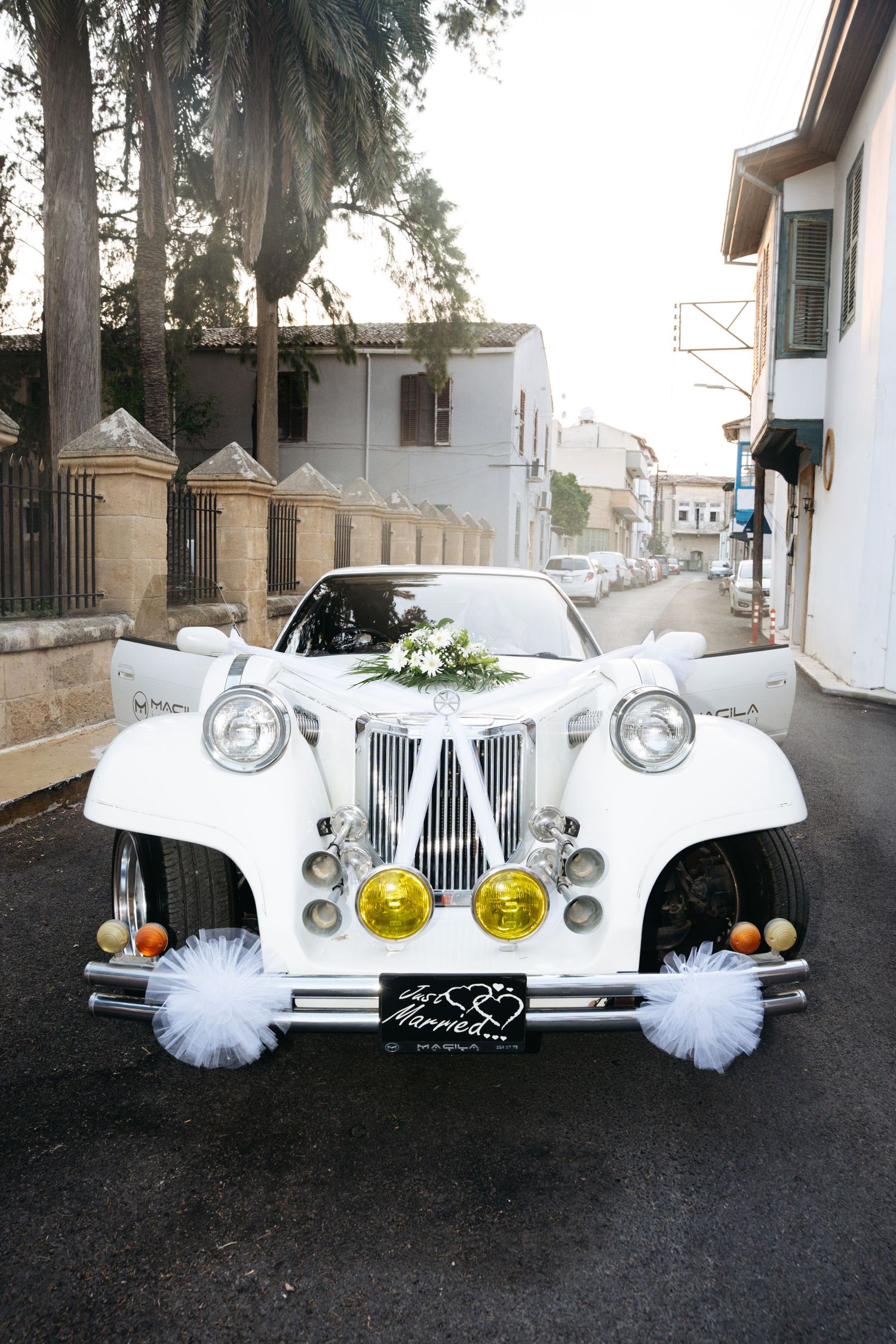 White vintage wedding car decorated with tulle bows, yellow headlights, and floral bouquet, parked on a quiet street.