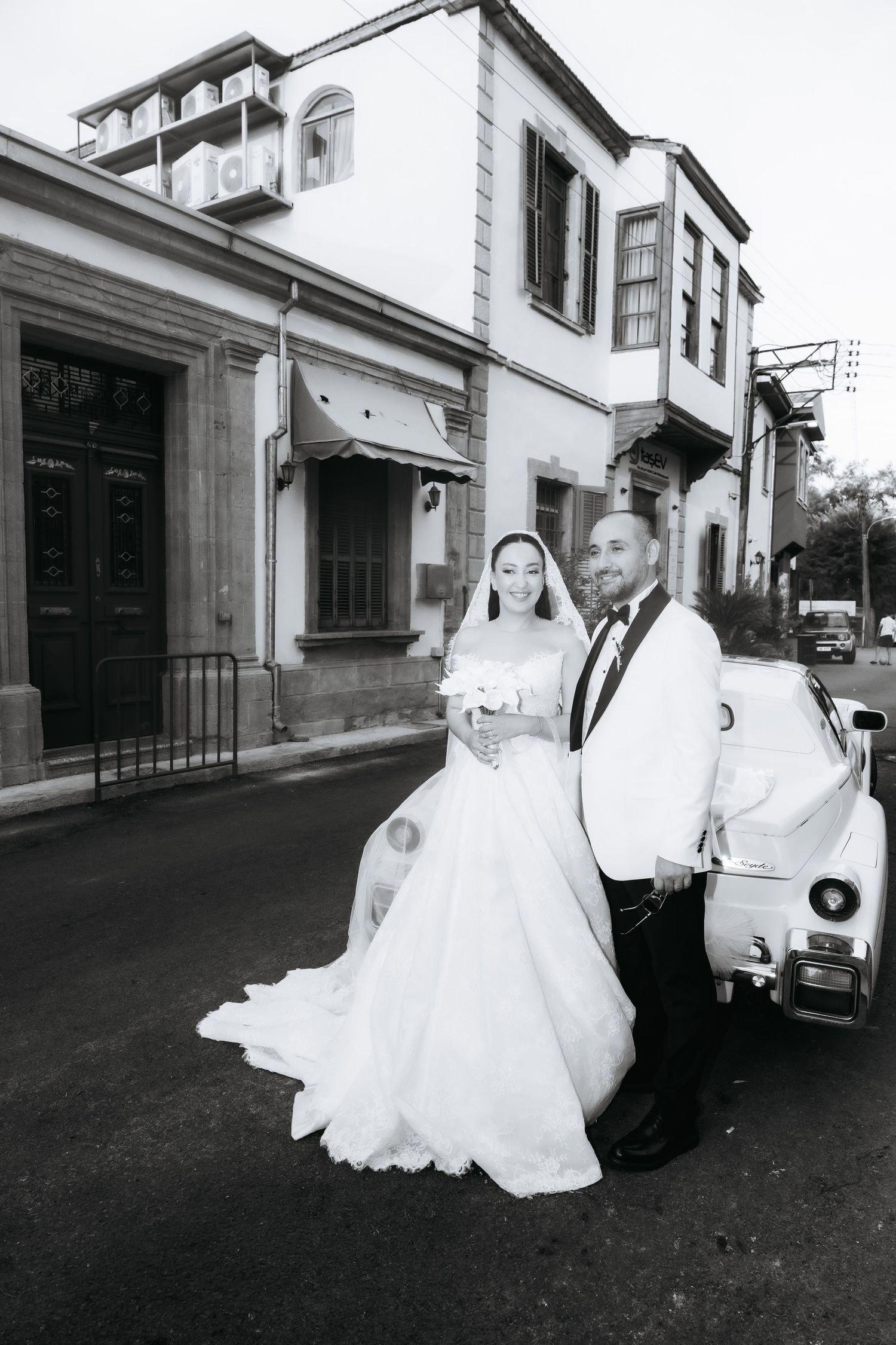 Black and white wedding photo of bride and groom posing beside a vintage white car on a charming cobblestone street.