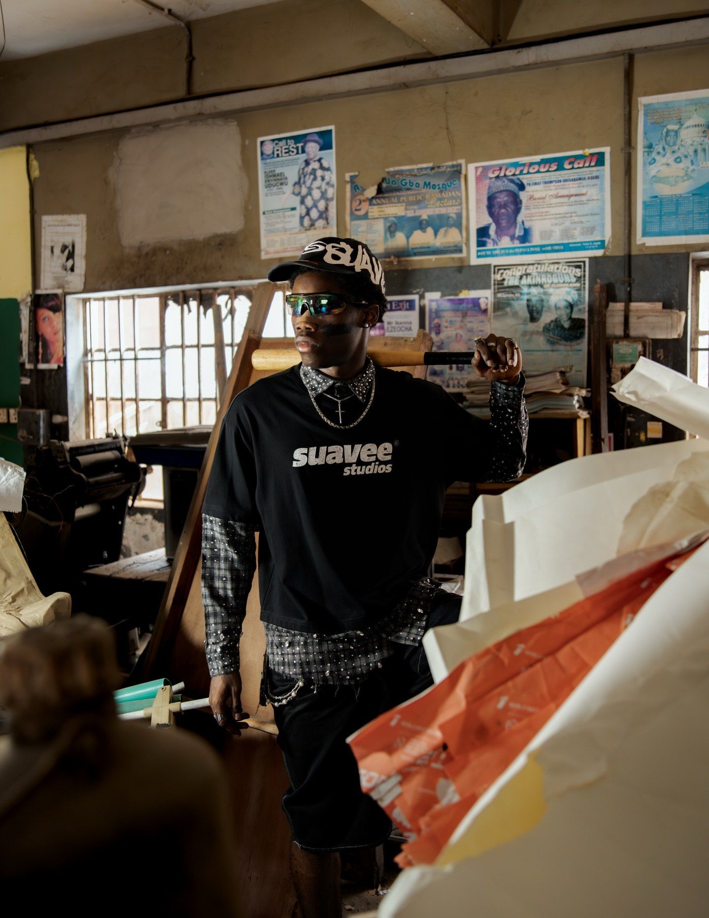 Person wearing Suavee Studios black sweatshirt and cap standing in a workshop surrounded by materials and posters.
