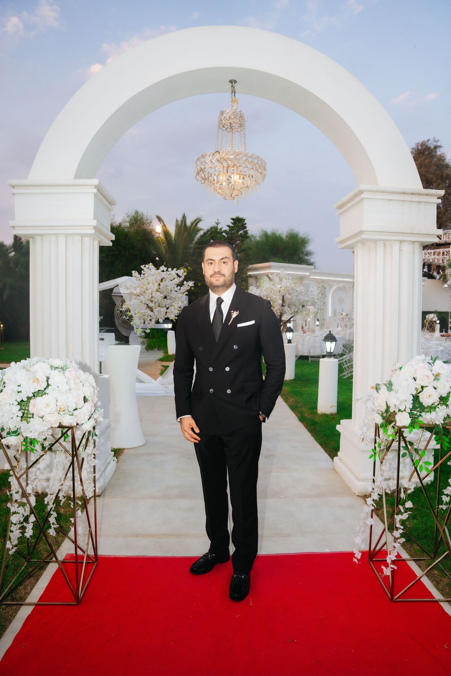 Groom in black double-breasted suit stands on red carpet under white arch with chandelier at outdoor wedding venue.
