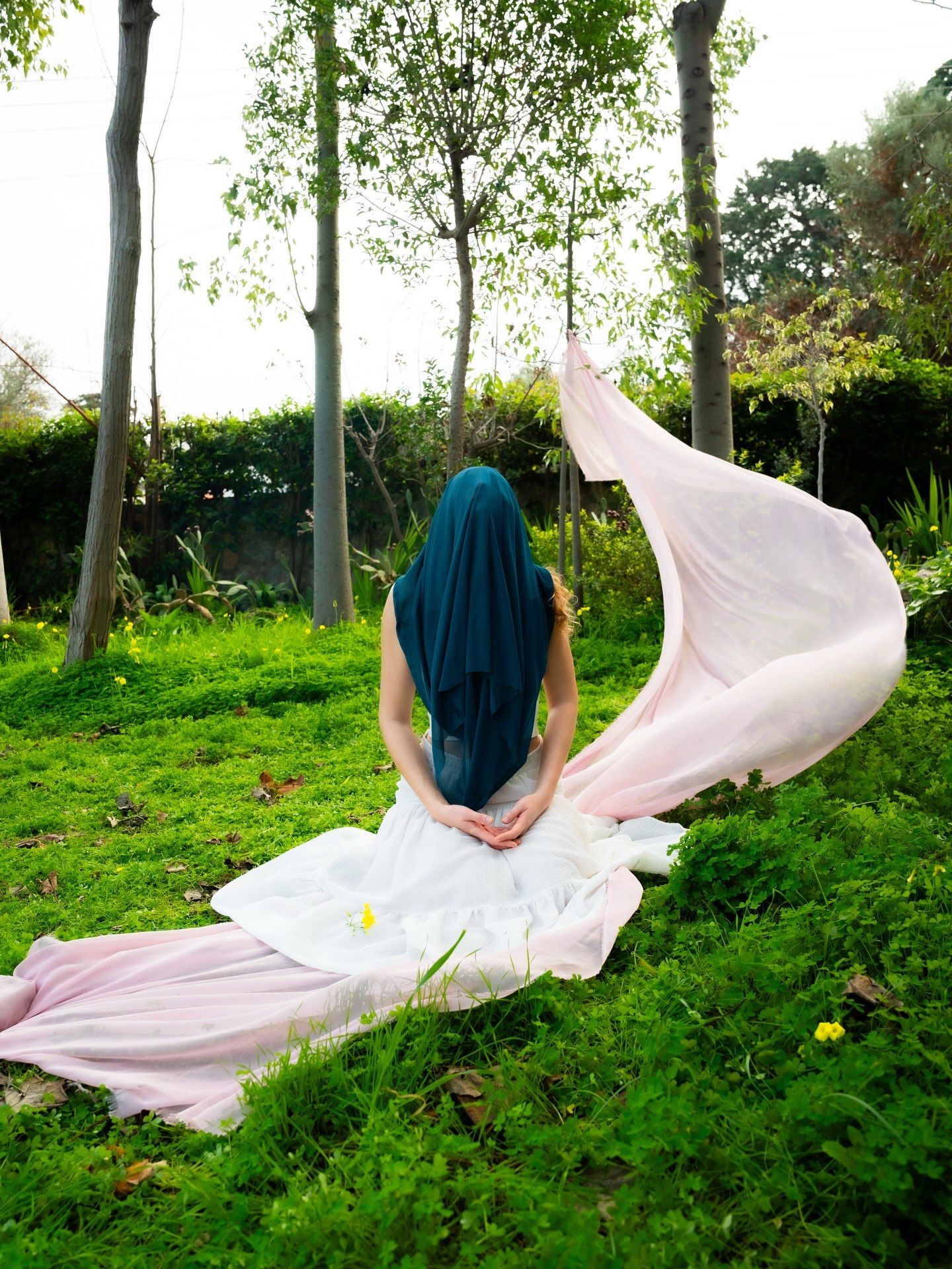 Woman in white dress with teal hair sitting on green grass with pink fabric flowing in the wind.