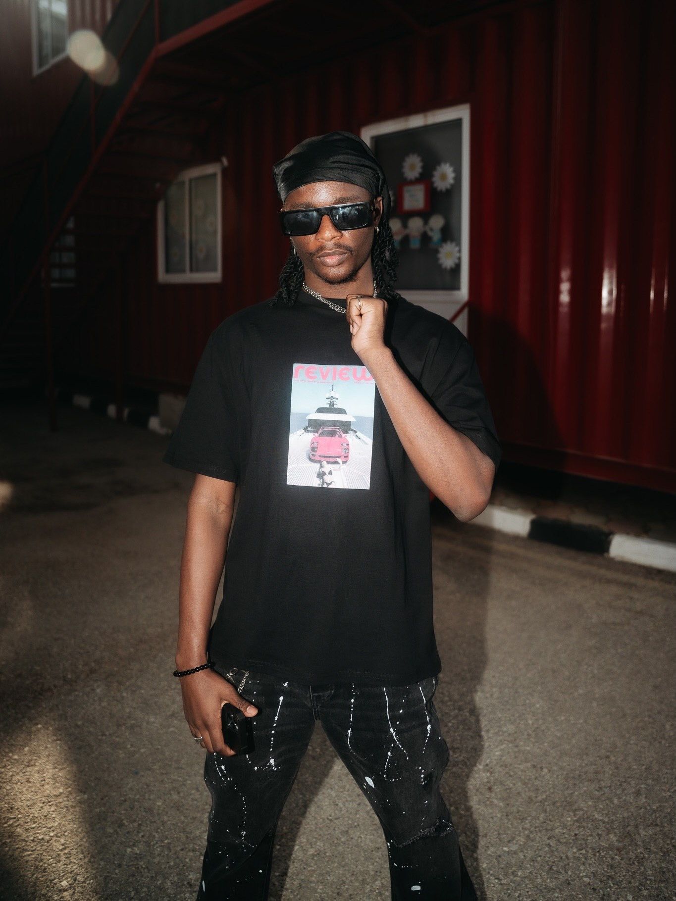 Young man in black graphic tee, sunglasses, and splatter-paint pants posing confidently at night outdoors.