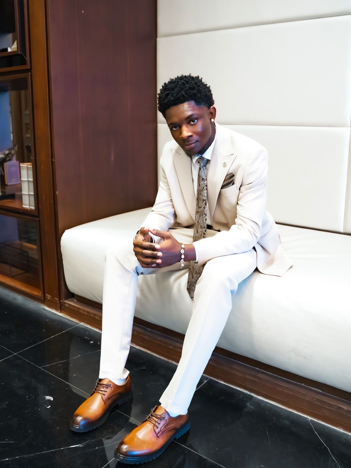 Stylish young man in cream suit with paisley tie and brown dress shoes sits on white bench.