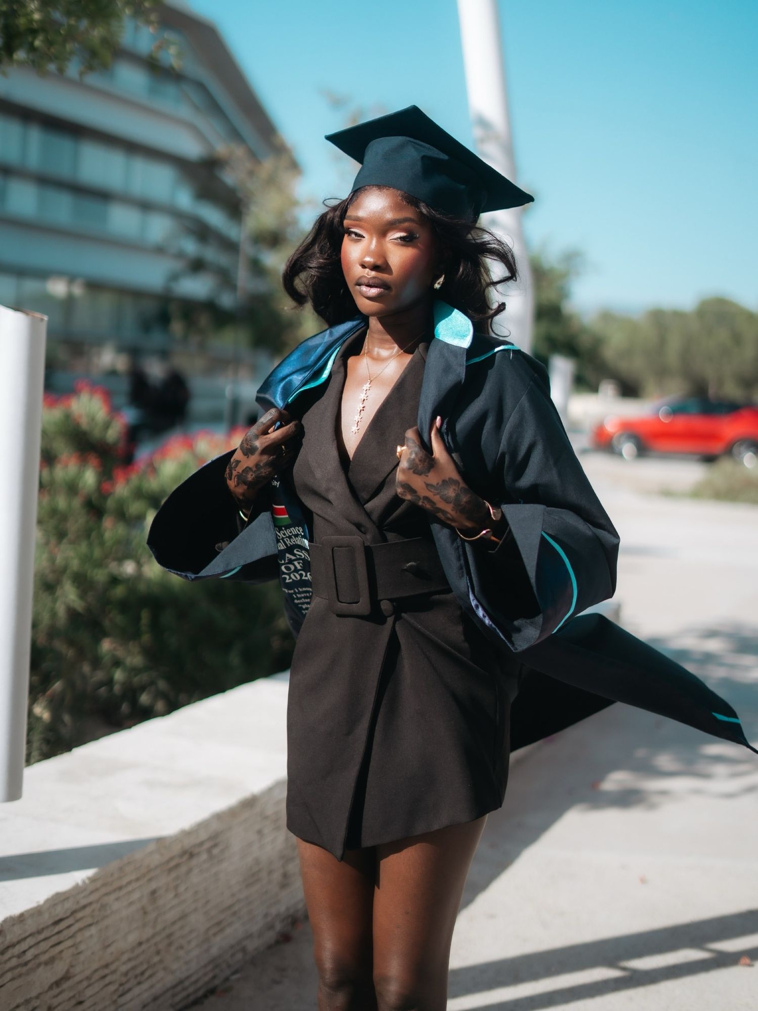 Young woman in black belted dress and graduation cap holds open academic gown outdoors on sunny day.