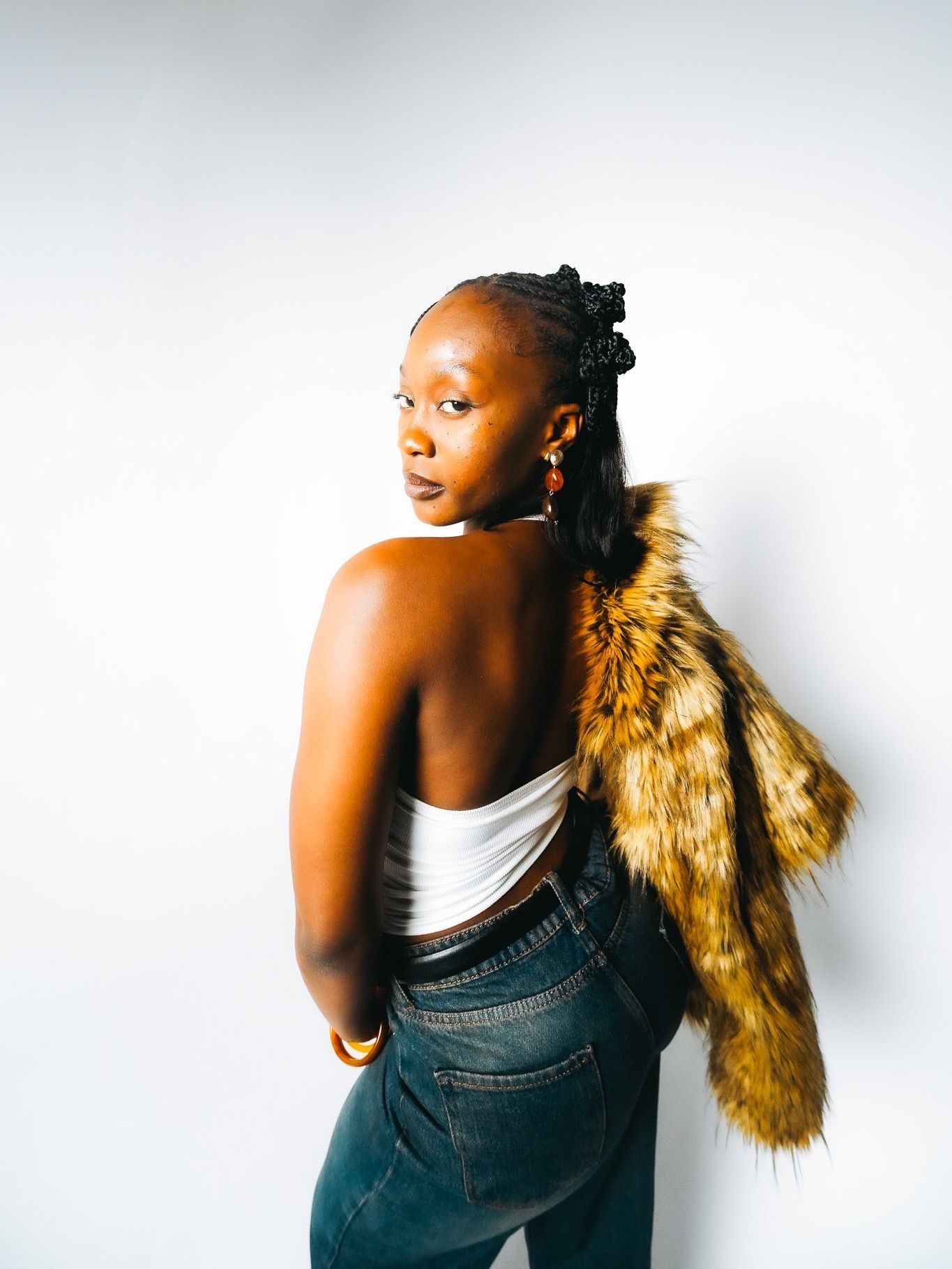 Black woman glances back over shoulder wearing faux fur coat draped over white crop top and jeans.