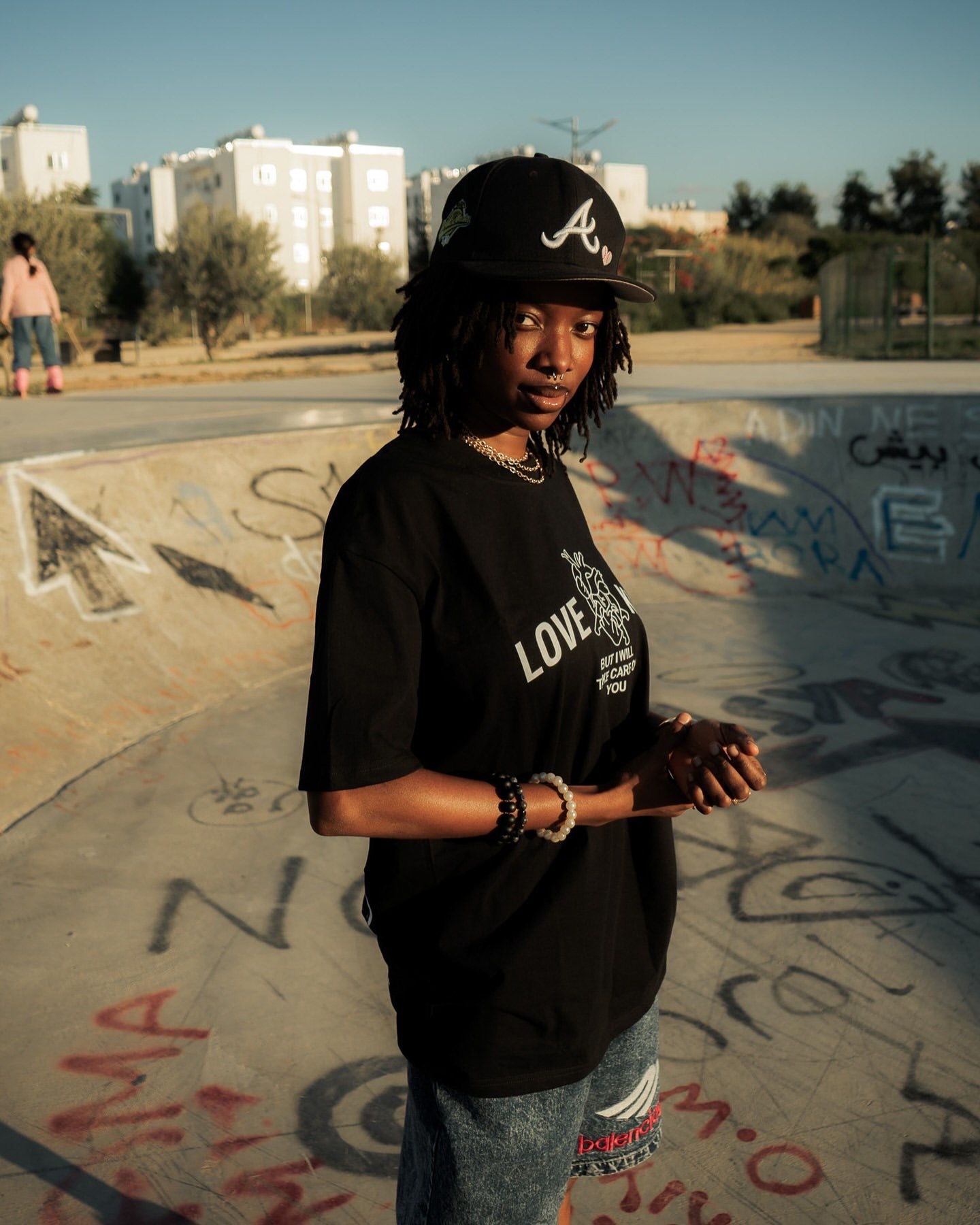 Young woman in black Atlanta Braves cap and graphic tee poses at a graffiti-covered skate park.