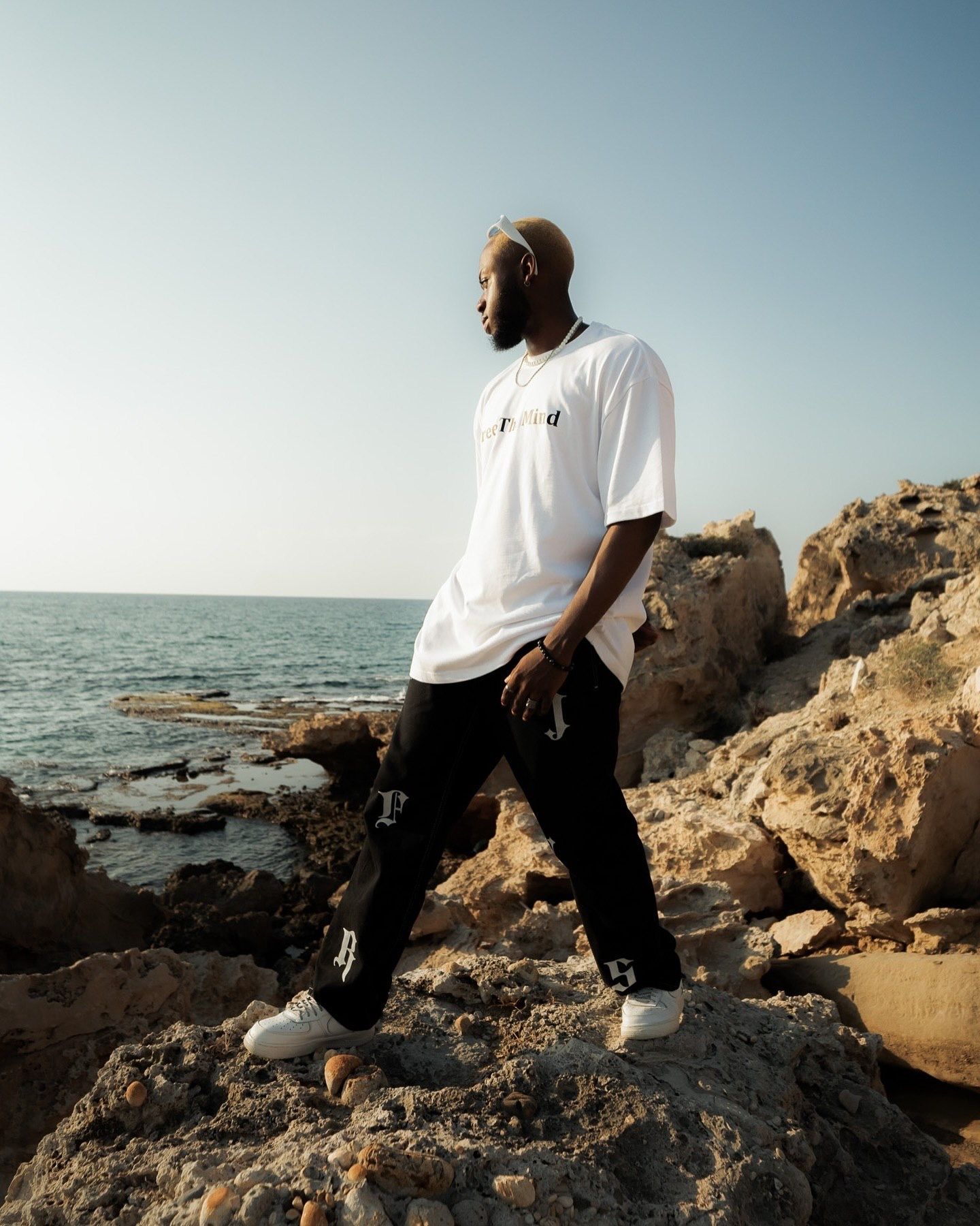 A young Black man in a white t-shirt and black pants stands on rocky coastal cliffs with the ocean in the background.