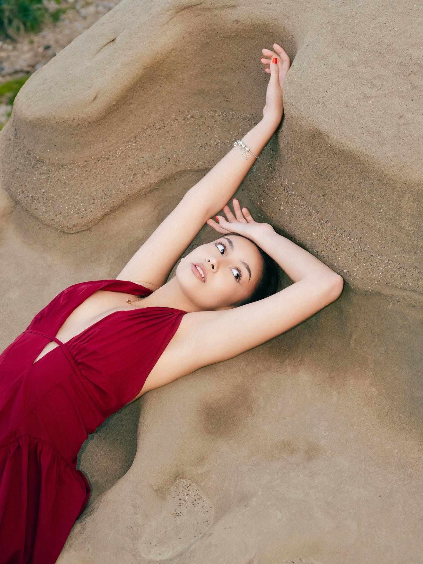 Woman in red dress posed gracefully against sandy rock formation outdoors.