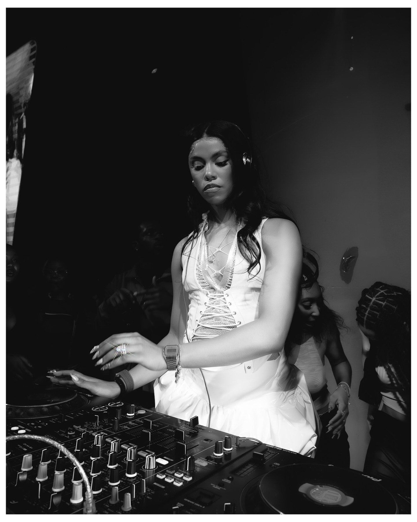 Black and white photo of a female DJ wearing headphones and a white dress, mixing at a DJ booth in a club.