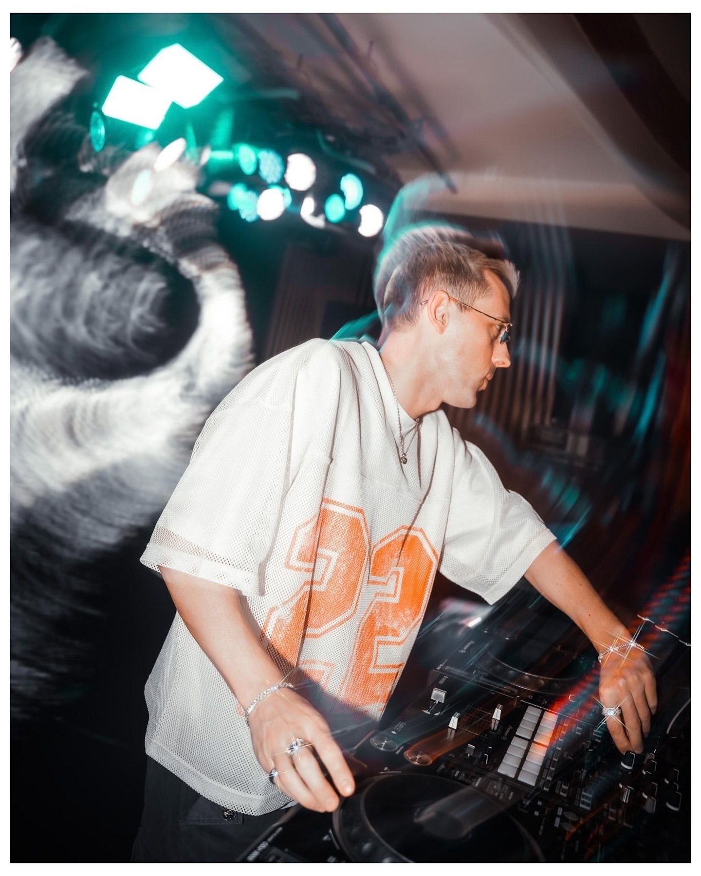 DJ in white jersey mixing tracks on turntables under colorful stage lights at a nightclub event.