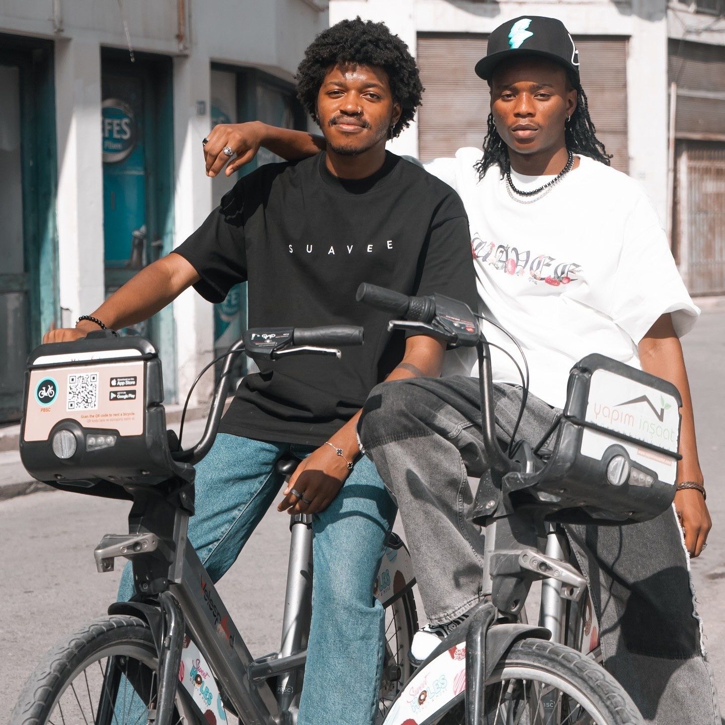 Two young men posing on bicycles on an urban street, wearing stylish streetwear outfits.