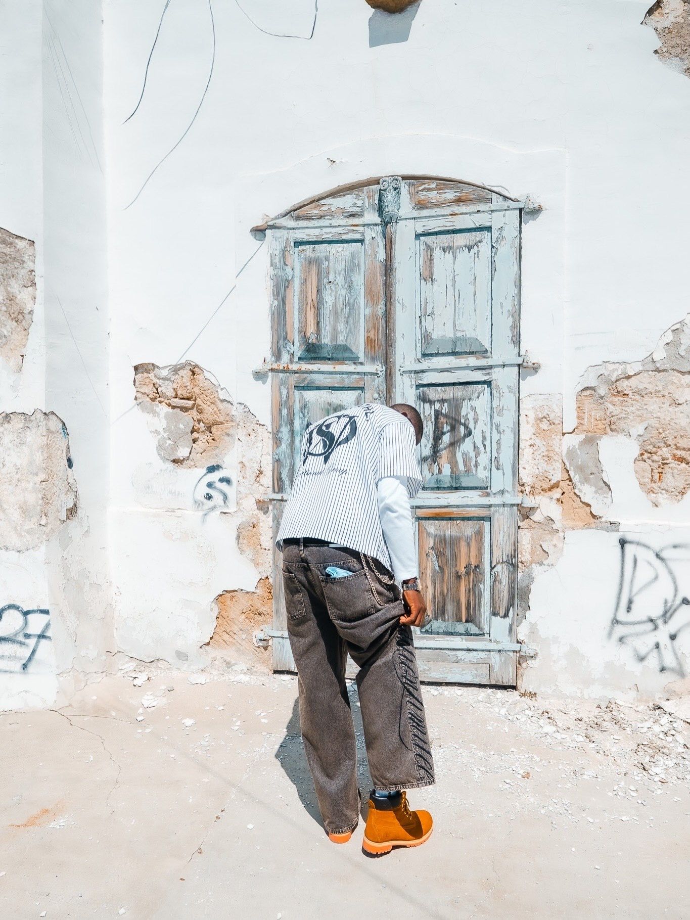 Person in gray hoodie and brown pants with orange shoes standing before a weathered white wall with an old teal door.
