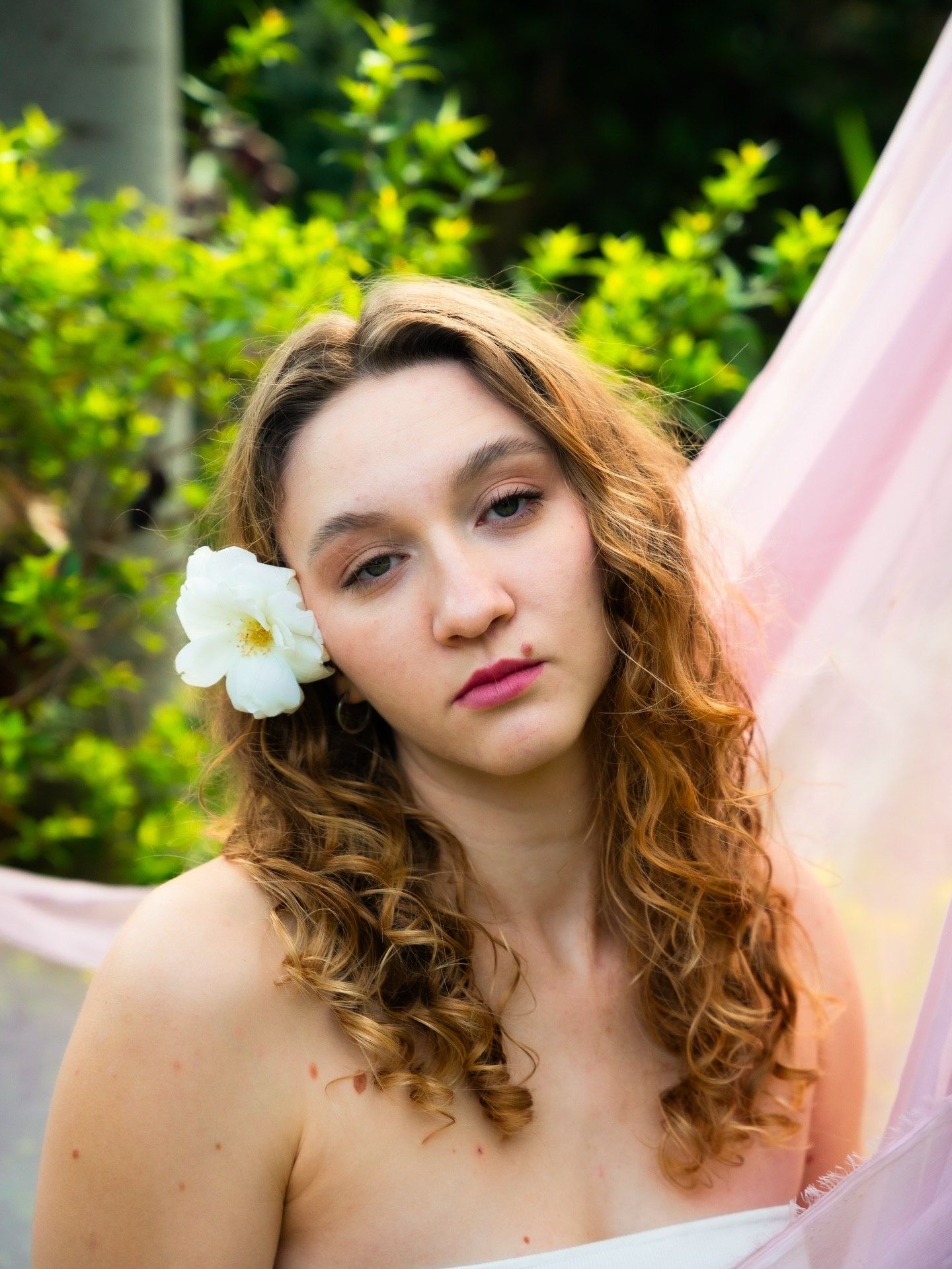 Young woman with curly auburn hair and white flower accessory poses outdoors with pink fabric and green foliage backdrop.