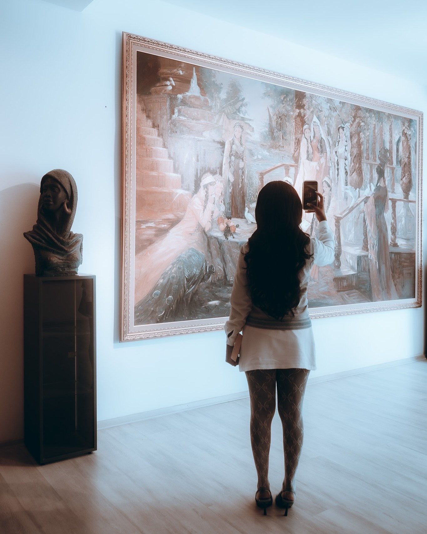Woman photographing a large classical painting in an art gallery beside a sculpted bust on display.