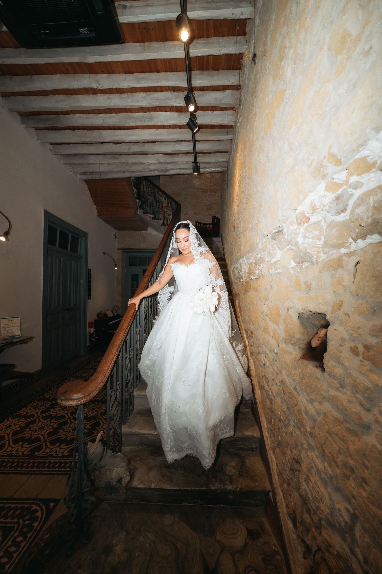 Bride in white ball gown and veil descends ornate staircase in rustic stone venue.