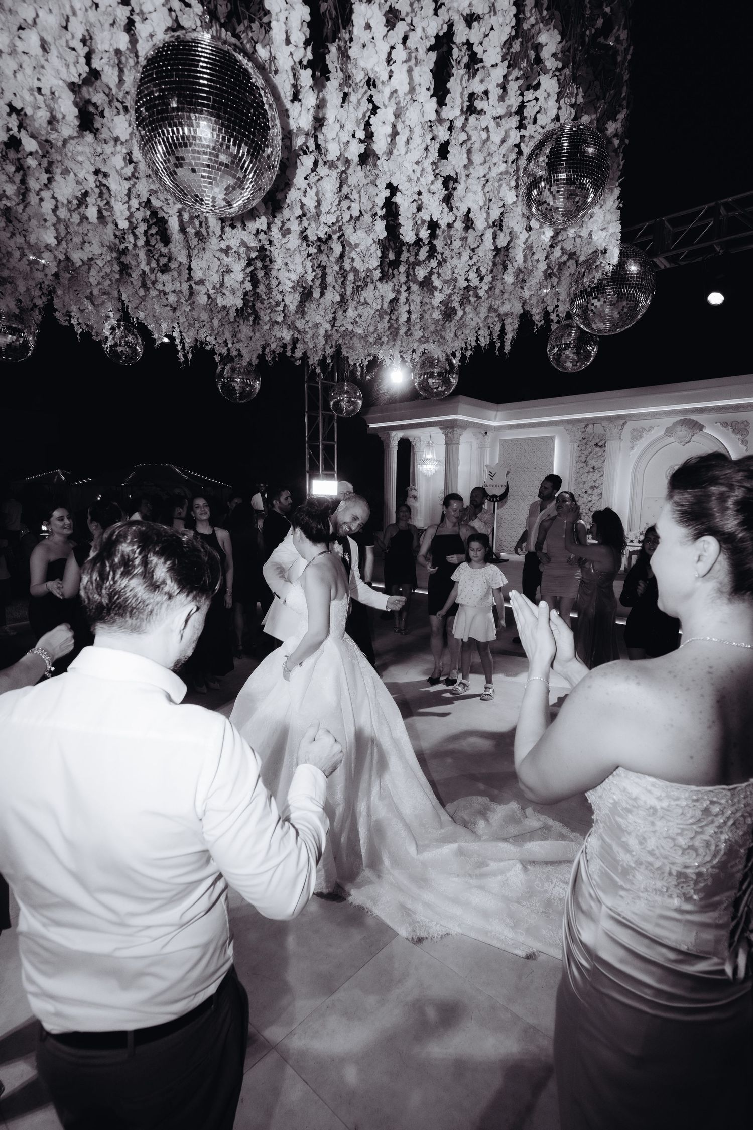 Black and white wedding reception photo with couple dancing under disco ball and floral ceiling decorations.