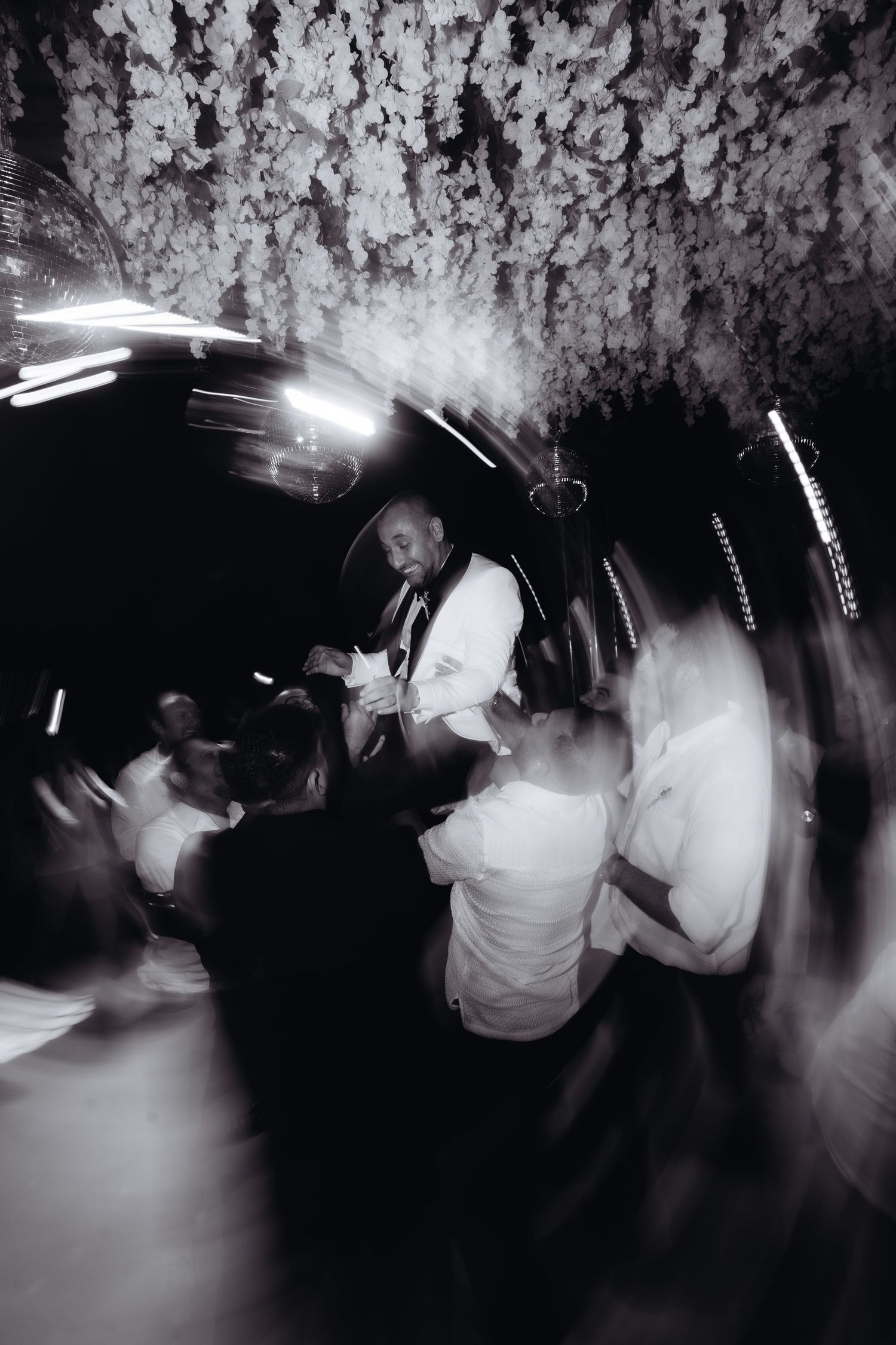 Black and white photo of wedding guests dancing energetically under floral ceiling decorations at a reception.
