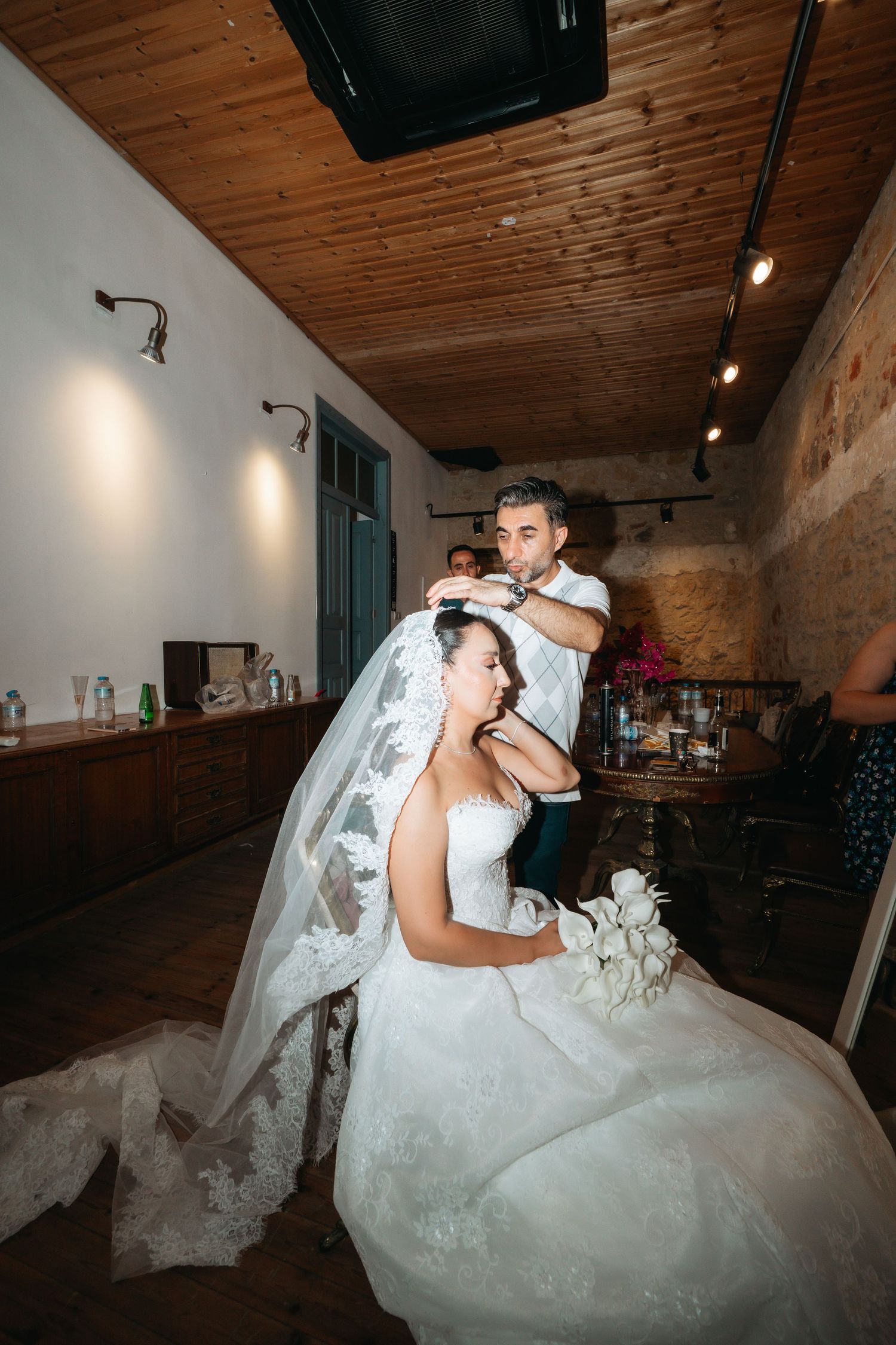 Groom placing veil on seated bride in white lace wedding dress inside rustic venue with wooden ceiling.