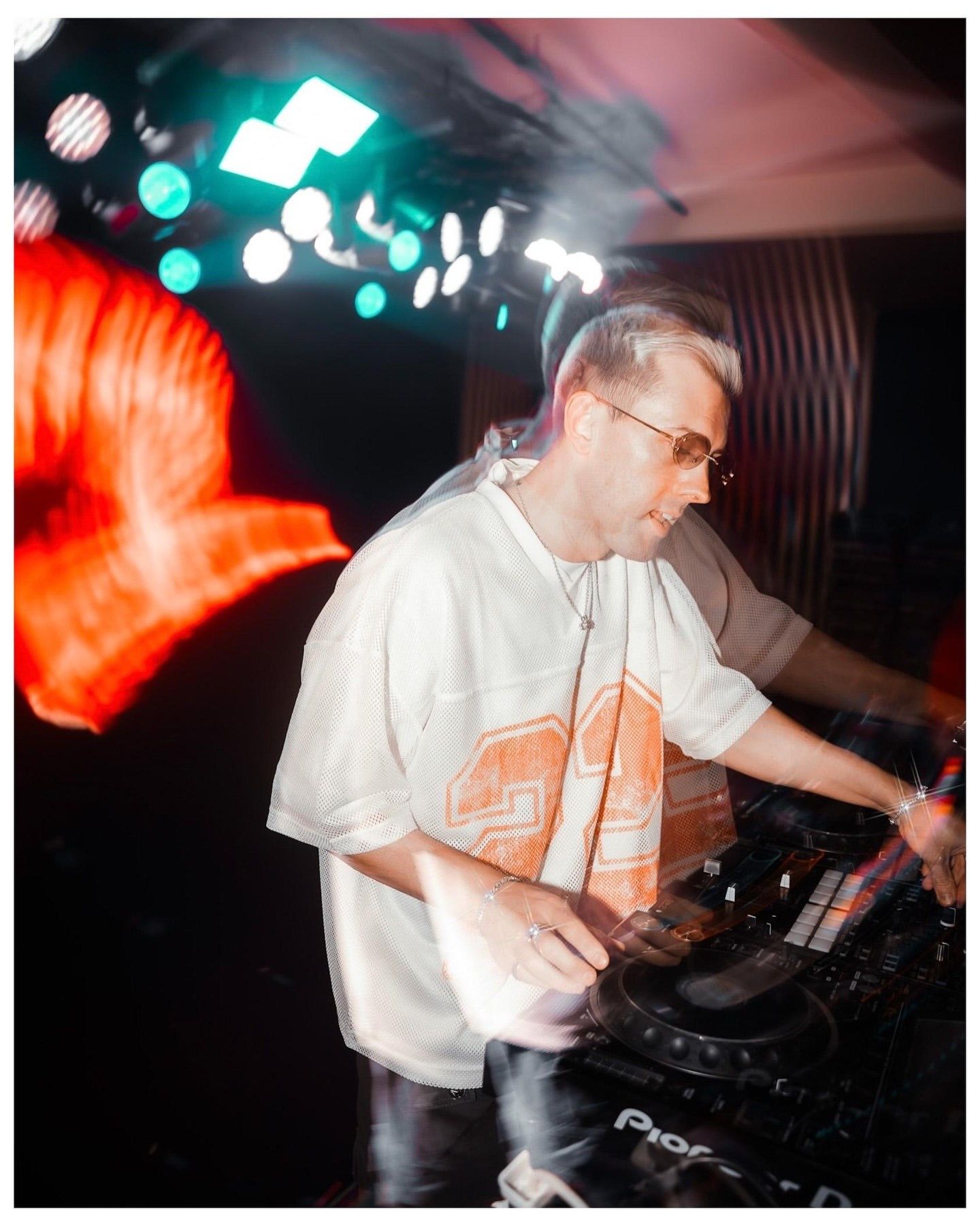 DJ performing at a nightclub with Pioneer decks, wearing a white oversized tee, illuminated by colorful stage lighting.