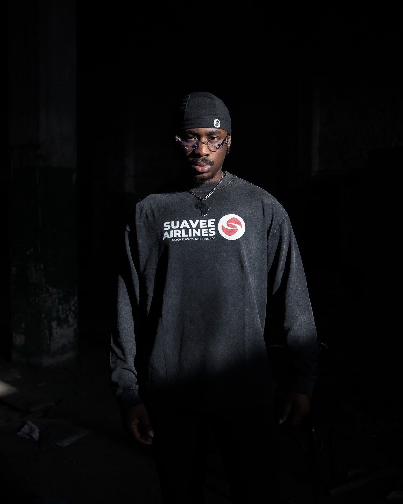 Man wearing a black Suavee Airlines sweatshirt and cap poses in dramatic low-key lighting.