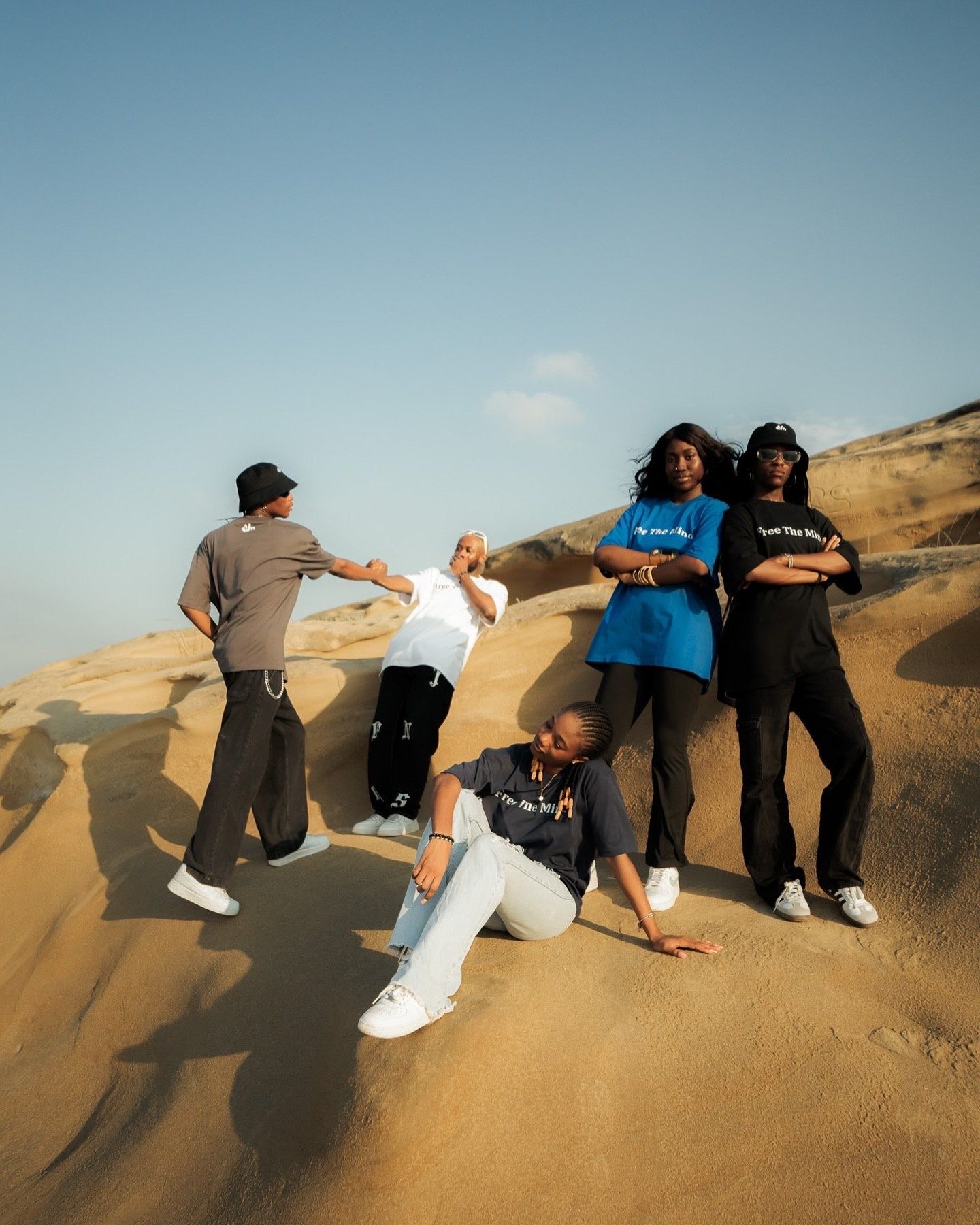 Five young people posing dynamically on golden sand dunes under a clear blue sky in casual streetwear.