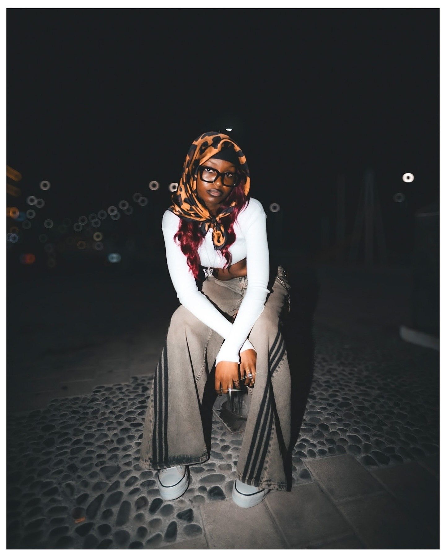 A young woman wearing a colorful hijab, white top, and wide-leg pants sits outdoors at night on a cobblestone surface.