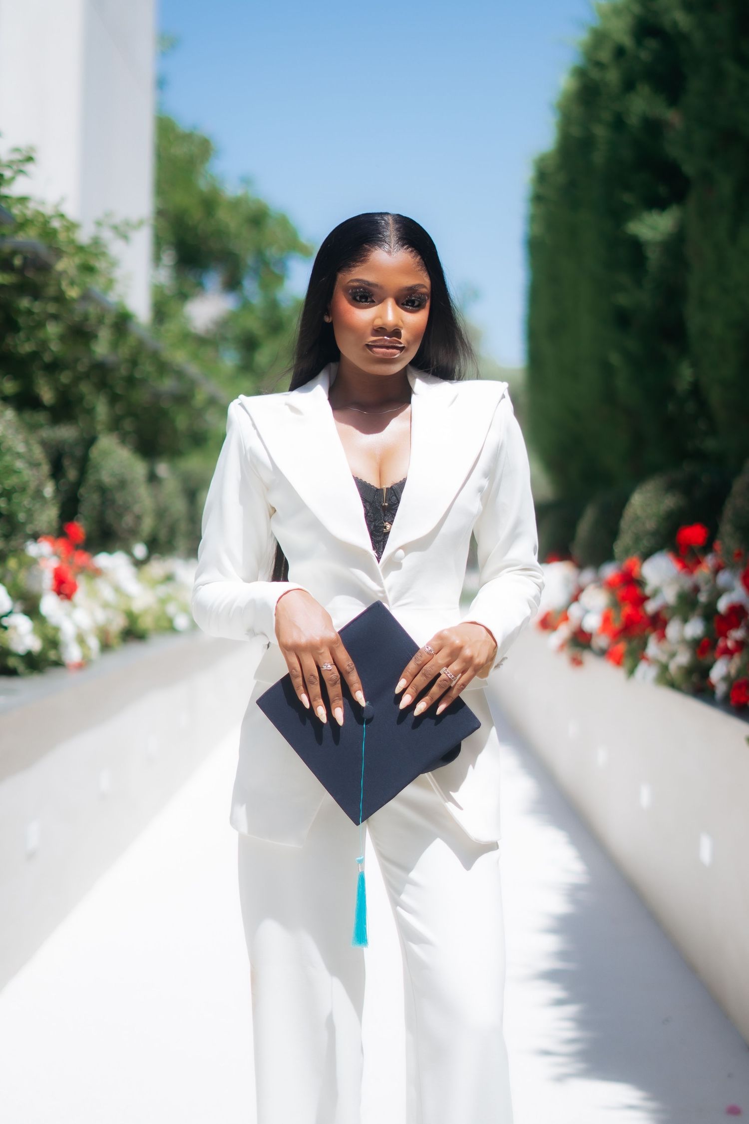 Confident Black woman in white suit holds navy graduation cap with teal tassel outdoors.