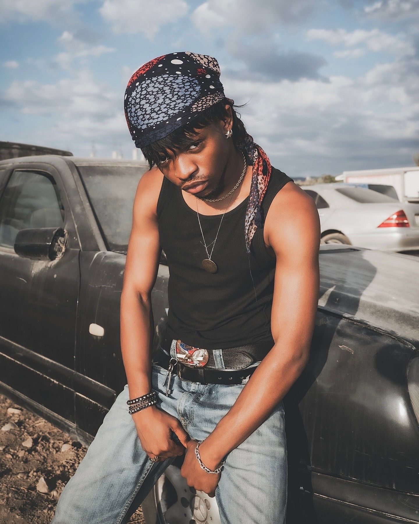 Young man in bandana, black tank top, and jeans poses near vintage cars in a junkyard setting.