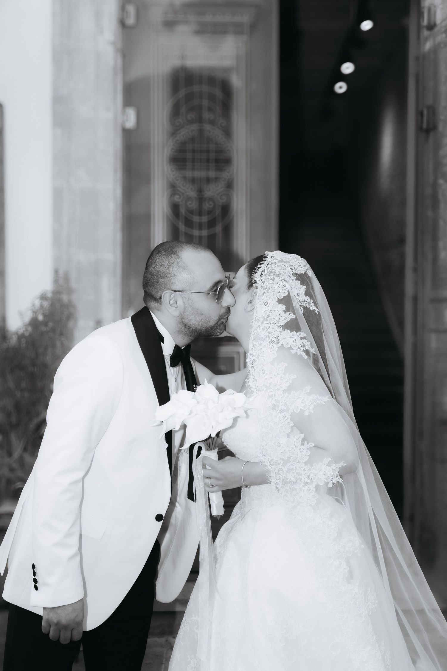 Black and white wedding photo of bride and groom sharing a kiss, bride in lace veil and gown, groom in white tuxedo.