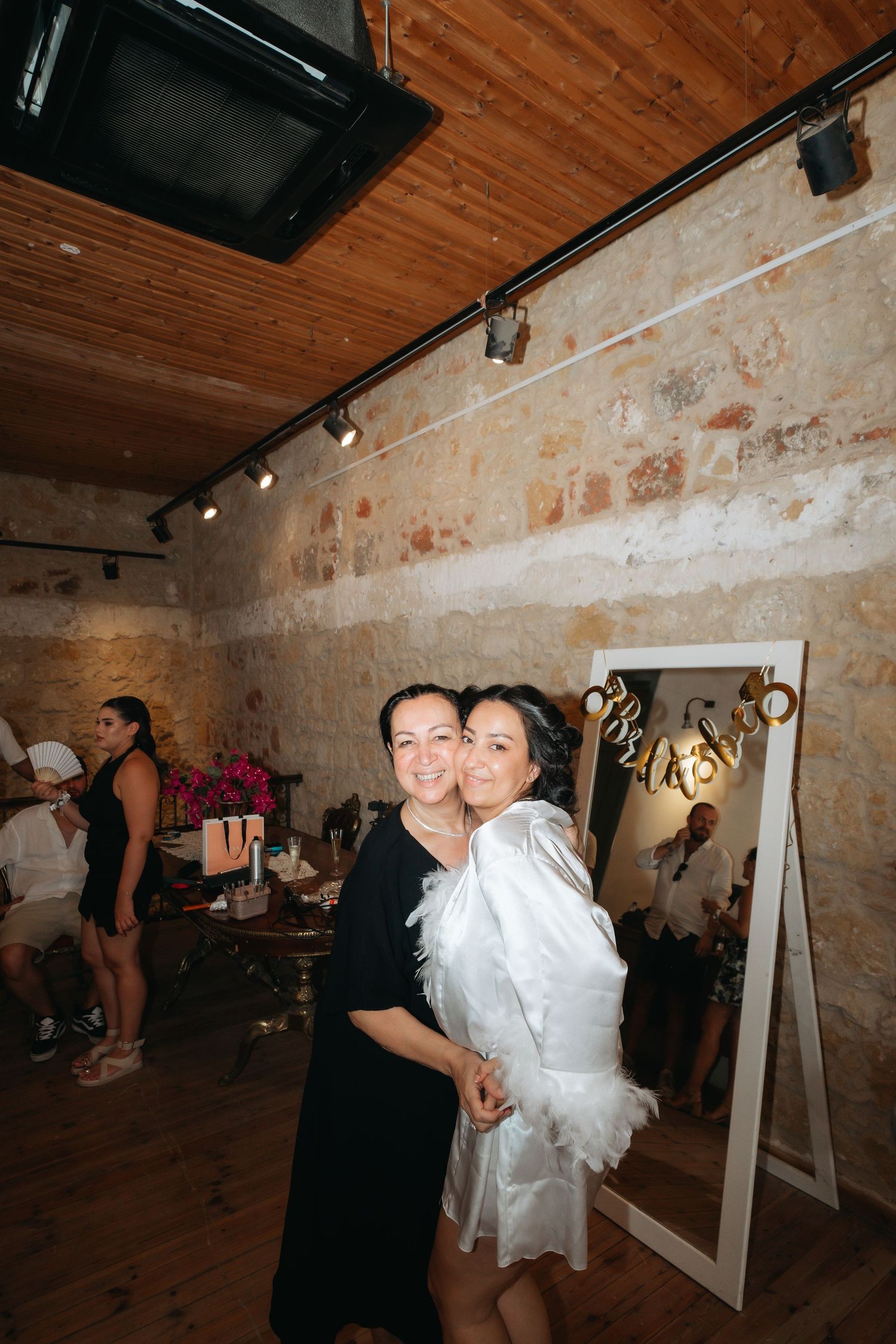 Two women smiling and posing together at an elegant stone-walled venue during a celebration event.