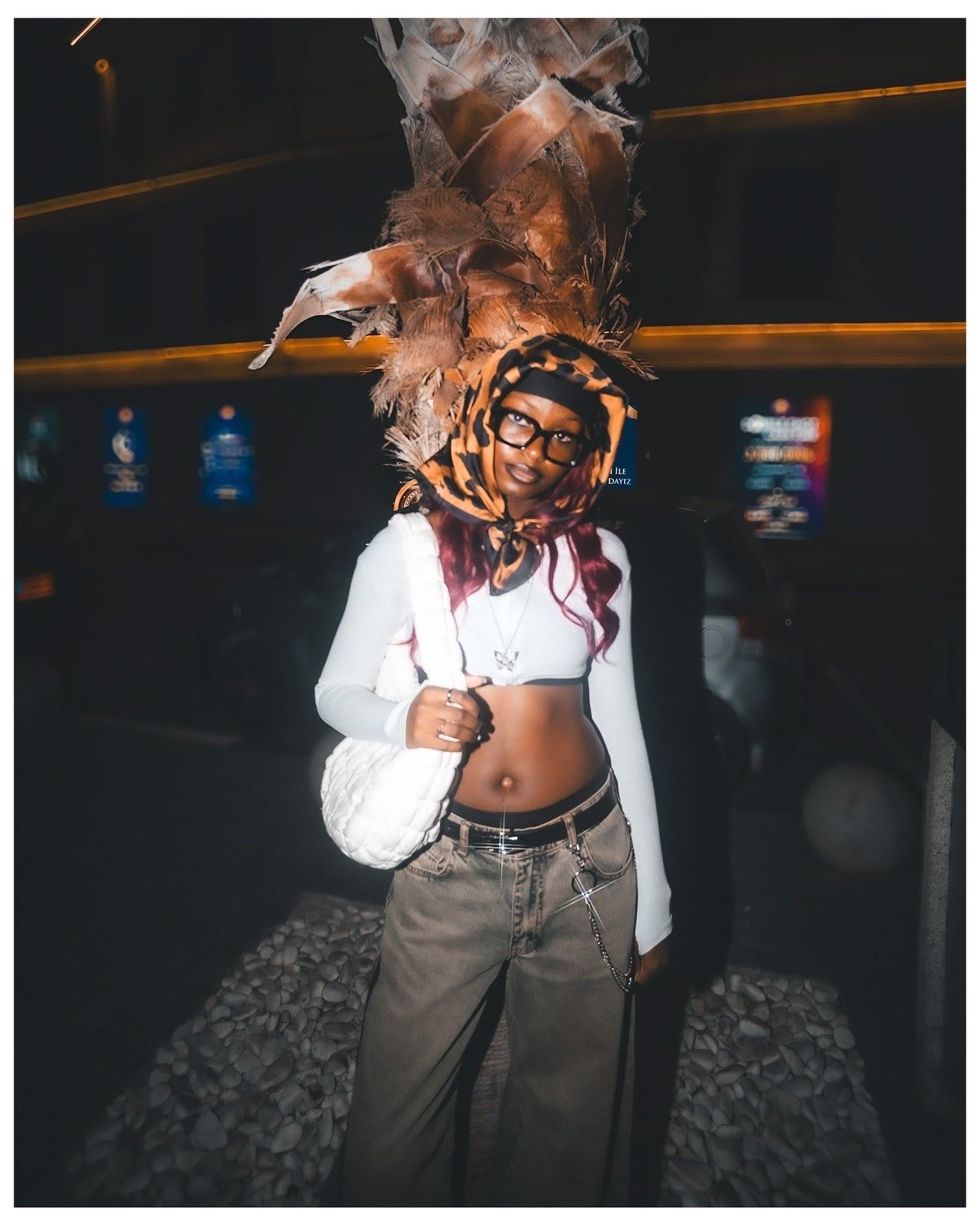 Person wearing a white crop top, glasses, and an elaborate feathered headdress poses indoors at a nighttime event.