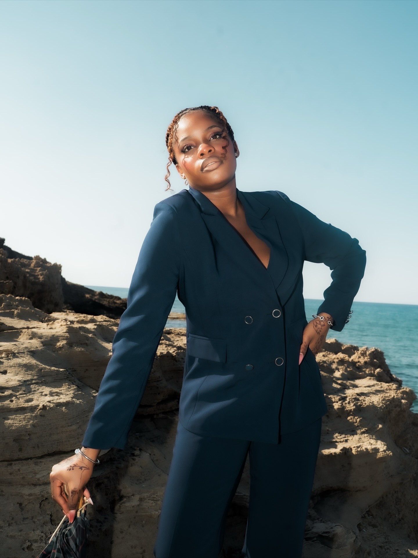 Woman in navy double-breasted pantsuit poses on rocky coastal cliffs with clear blue sky.