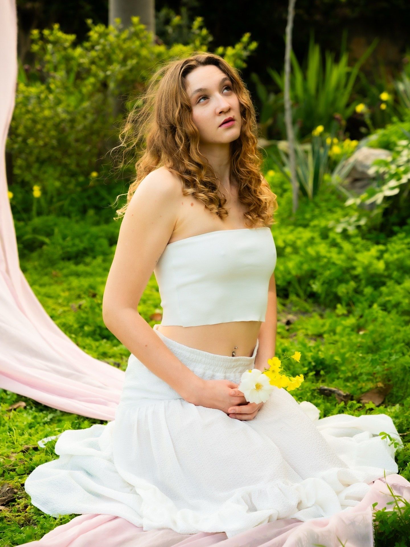 Young woman in white crop top and skirt holding a flower, sitting outdoors in a lush green garden setting.